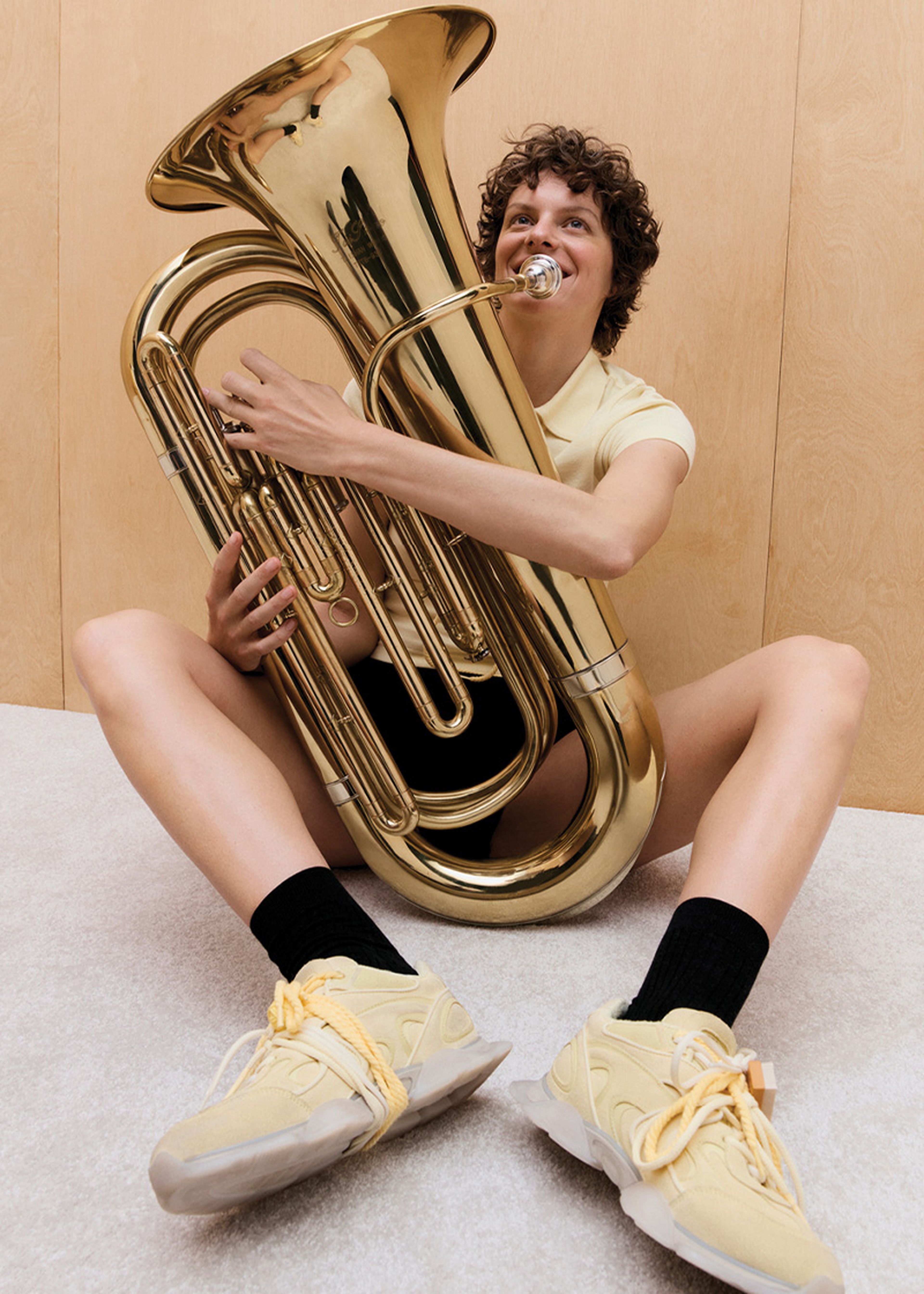 Person sitting on the floor, playing a large brass tuba, wearing a yellow shirt and Eris sneakers, in front of a wooden wall.