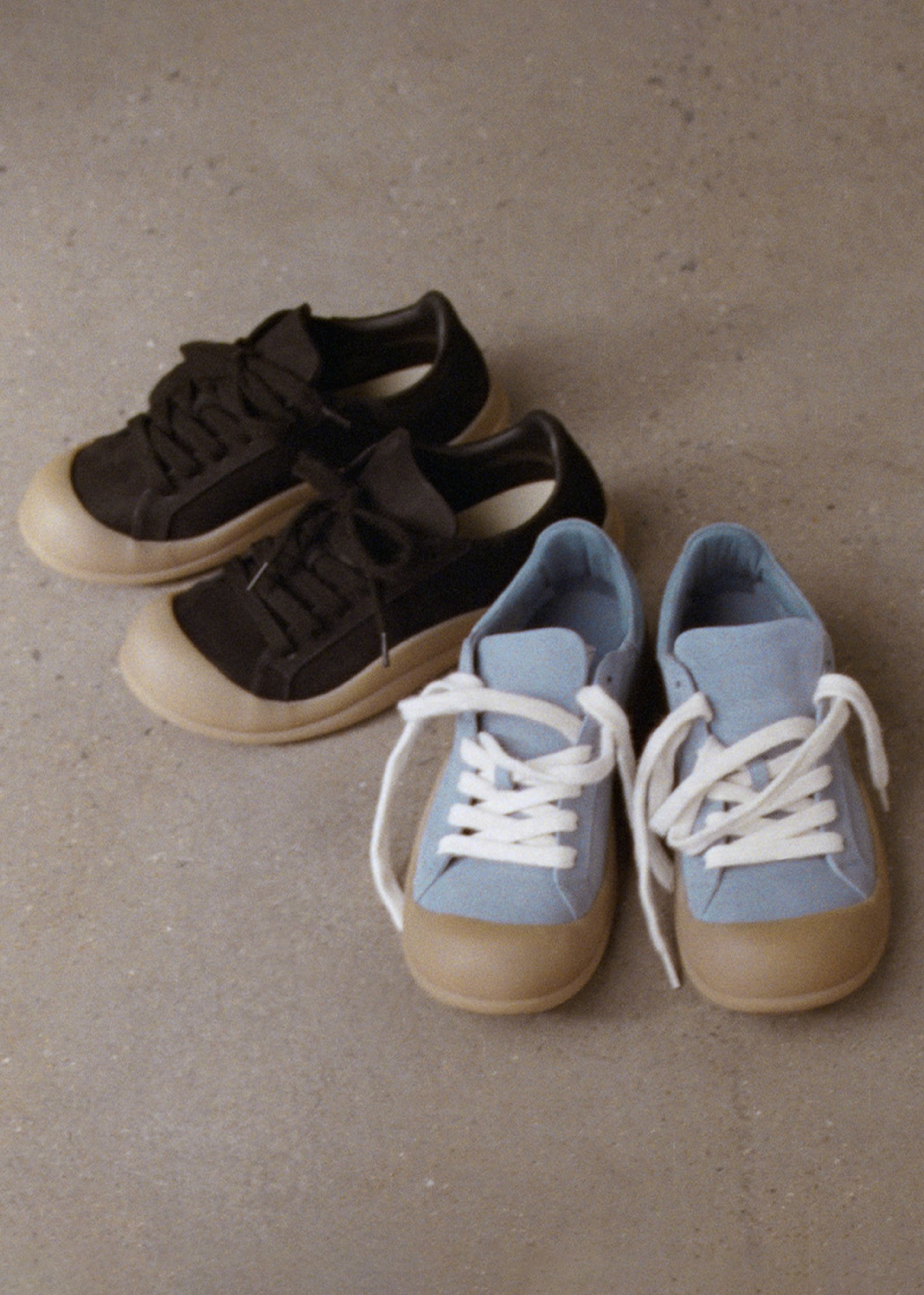 Two pairs of sneakers on a concrete floor: one pair is black with beige soles, the other is light blue with white laces and beige soles.