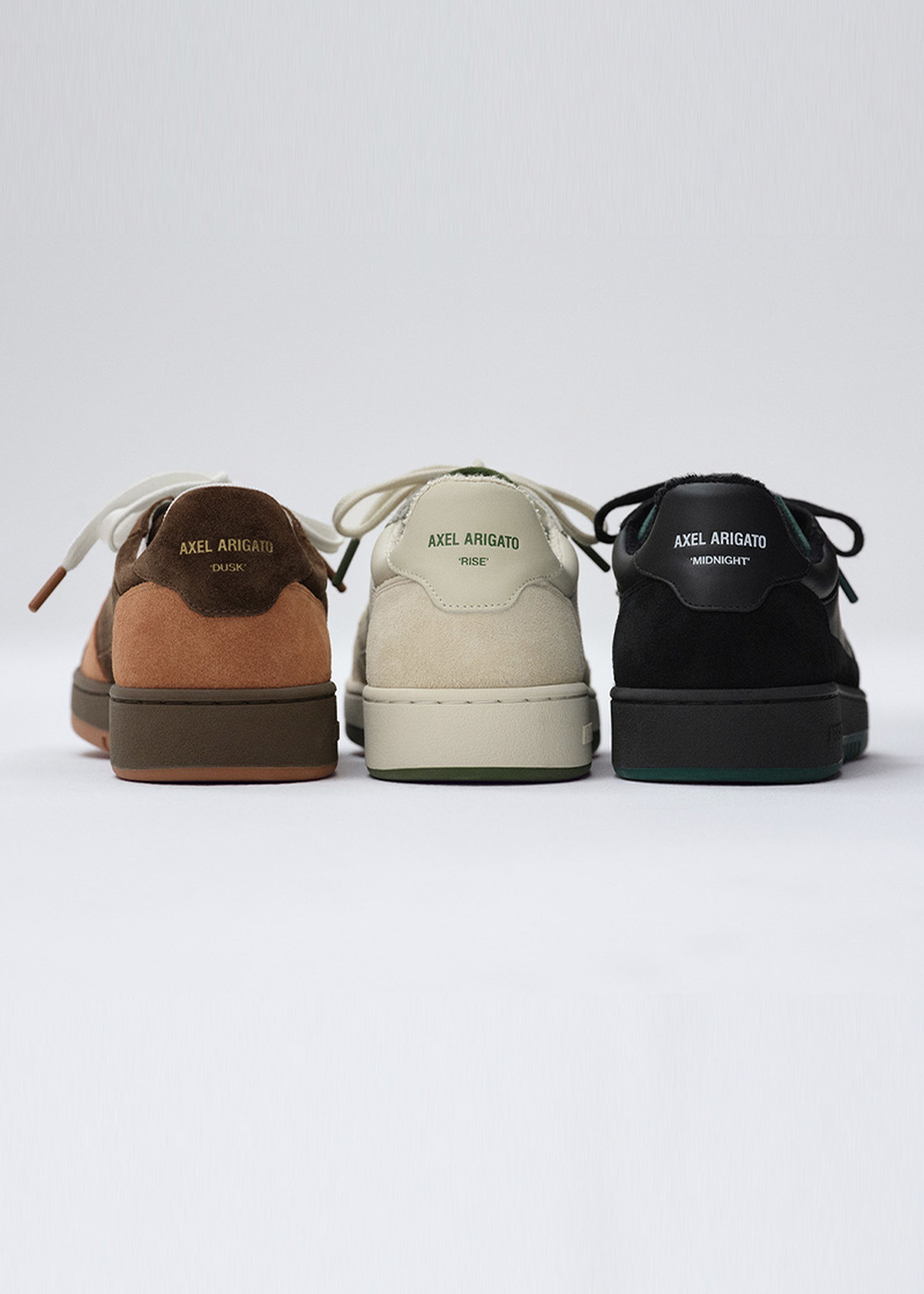 Three pairs of Axel Arigato sneakers in brown, beige, and black, viewed from the back against a plain background.