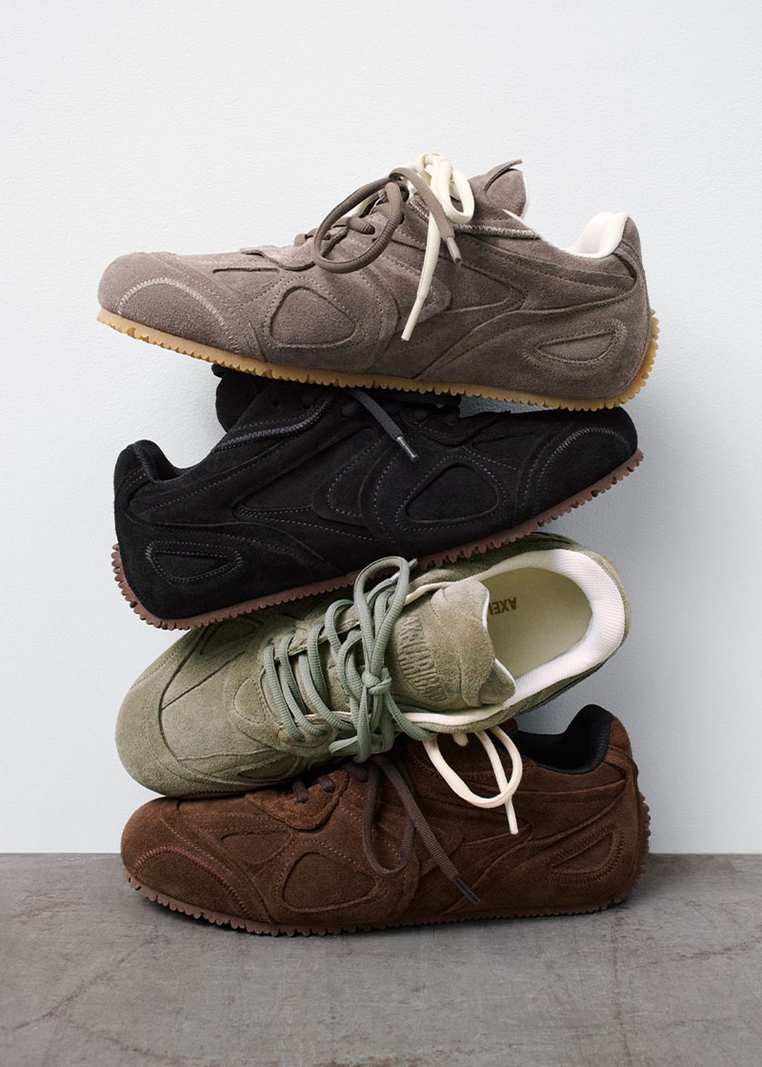 Four stacked sneakers in different colors: grey, black, green, and brown, against a plain background.