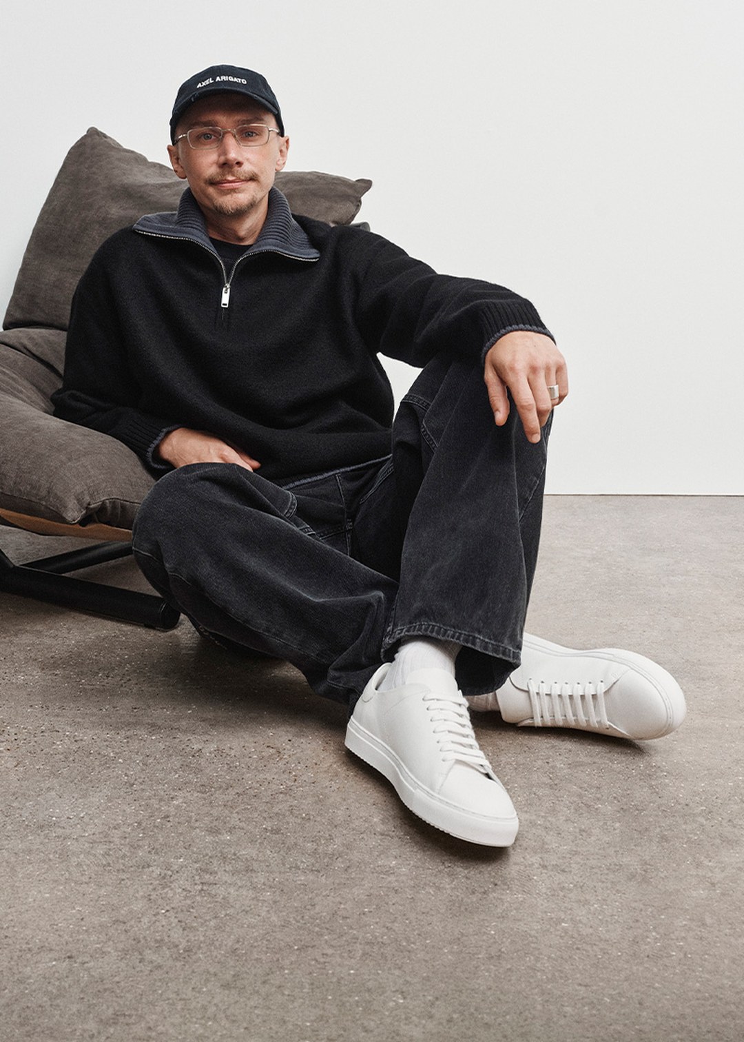 Man in a black sweater, jeans, and white sneakers, sitting on a cushioned chair with a relaxed pose, wearing a cap and glasses.