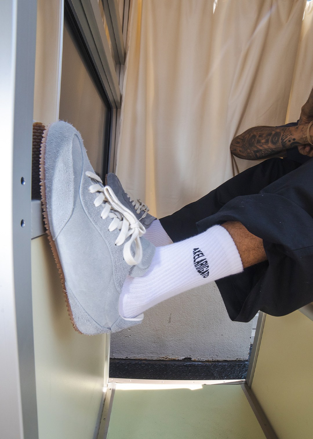 Person seated with legs outstretched, wearing gray suede sneakers, white socks, black pants, and displaying a tattooed arm in a brightly lit space.