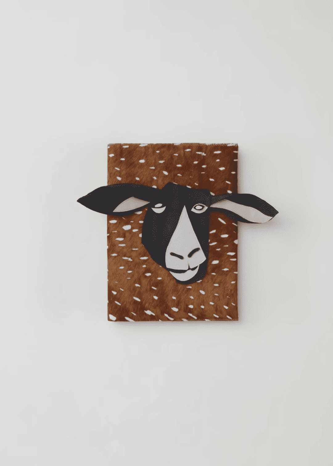 A wall-mounted, decorative goat head with black and white features protruding from a brown, speckled rectangular backing.