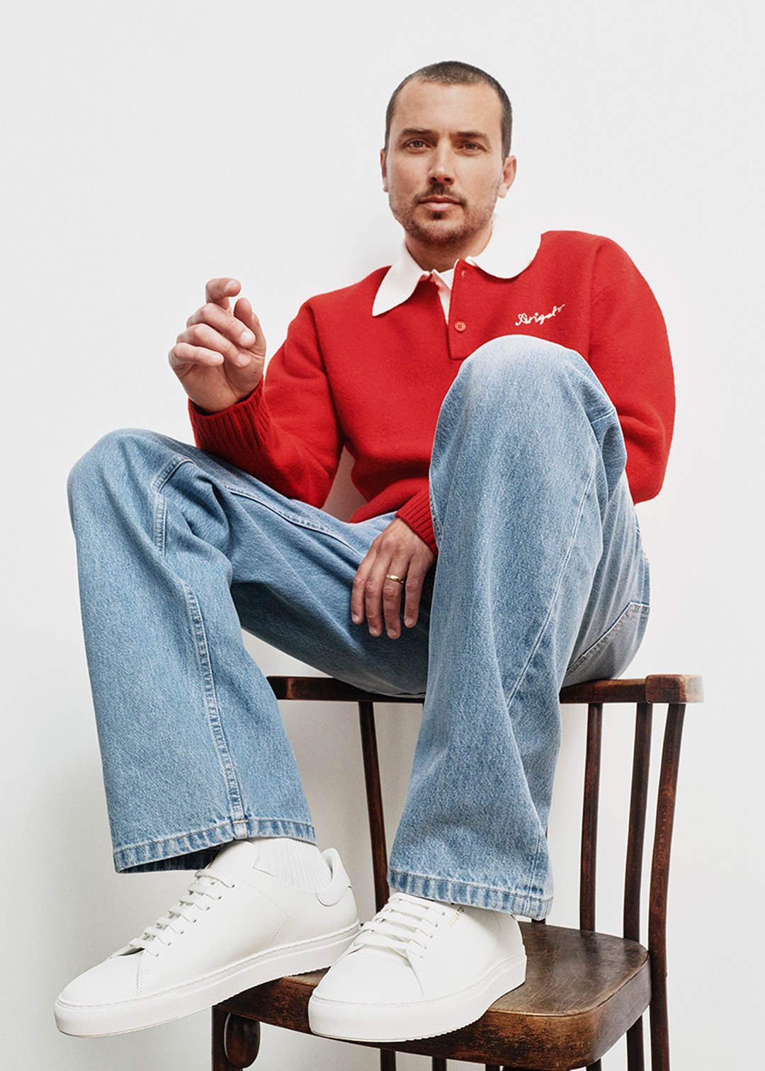 Man in red sweater with white collar and blue jeans sits on a chair, wearing white sneakers, in a relaxed pose against a plain background.