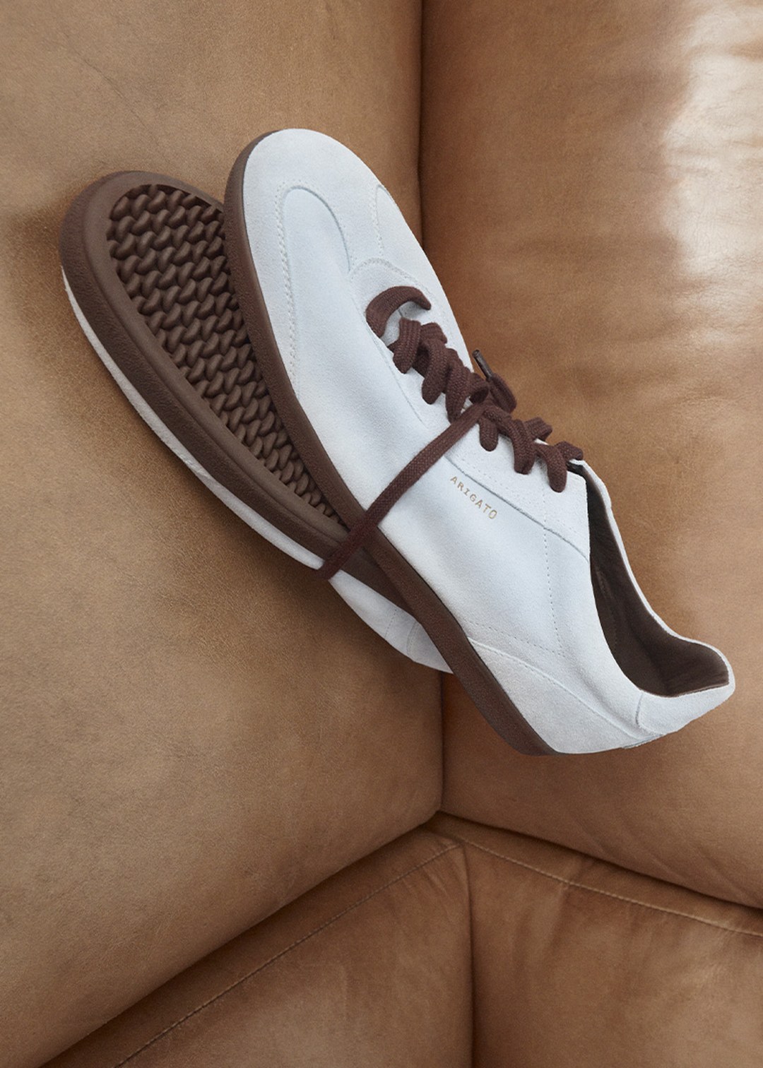White sneakers with brown soles and laces on a tan leather couch, featuring a textured waffle-patterned sole.