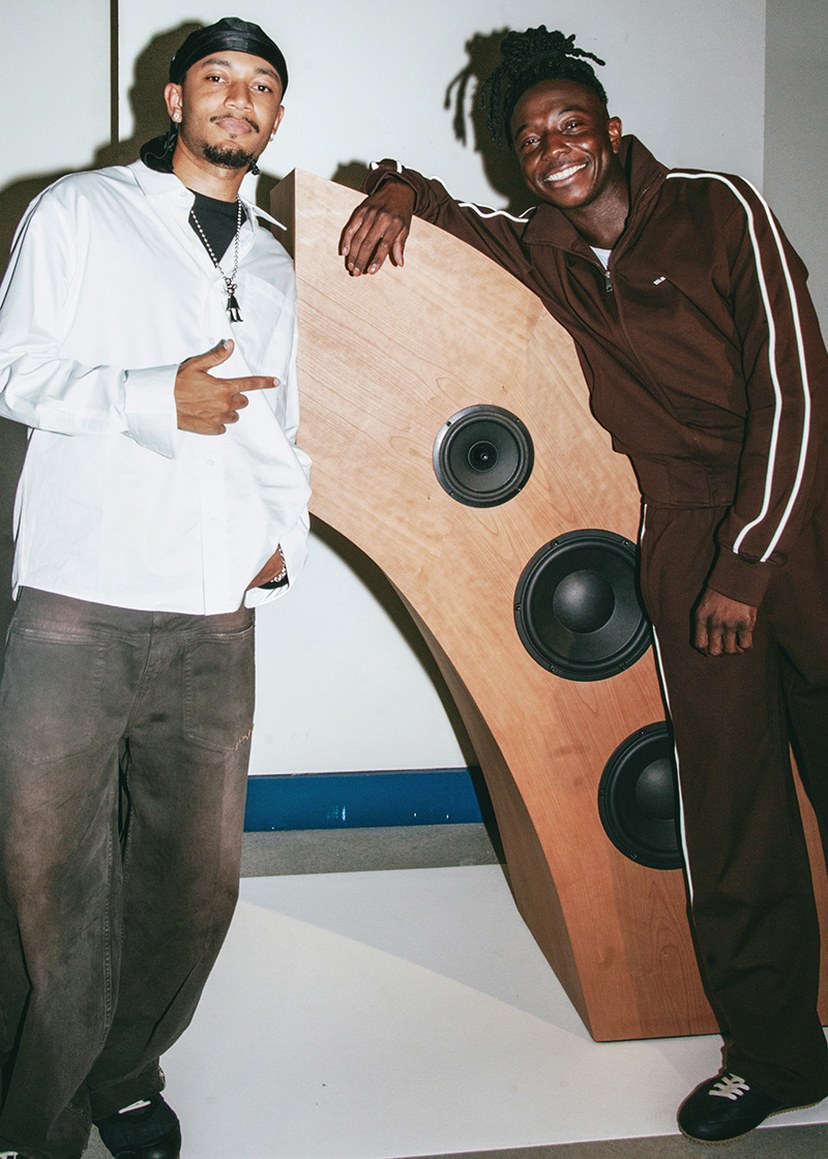 Two people smiling and posing beside a uniquely shaped wooden speaker with three circular speakers, inside a room.