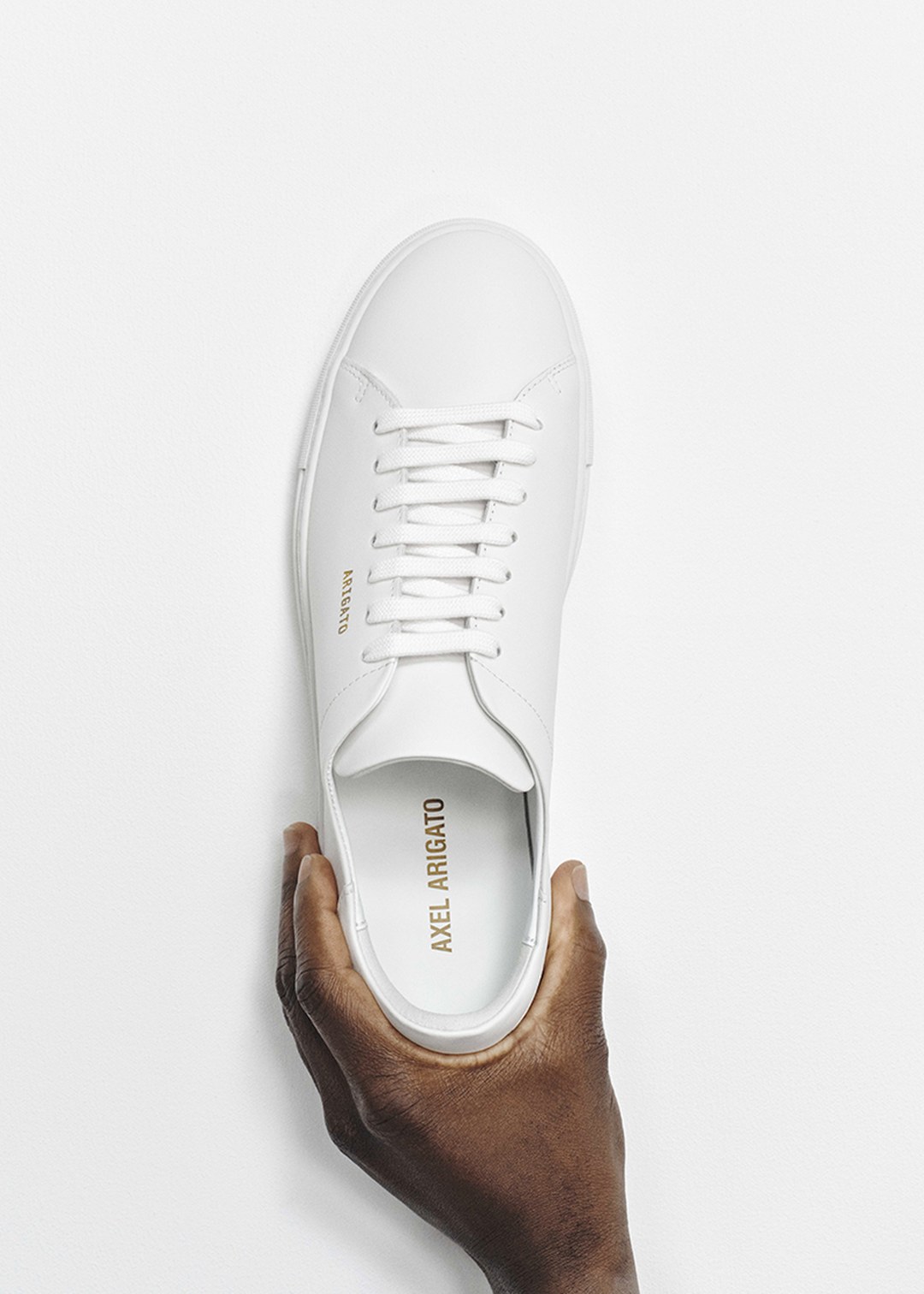 A hand holding a sleek, white sneaker with gold branding, viewed from above against a plain white background.