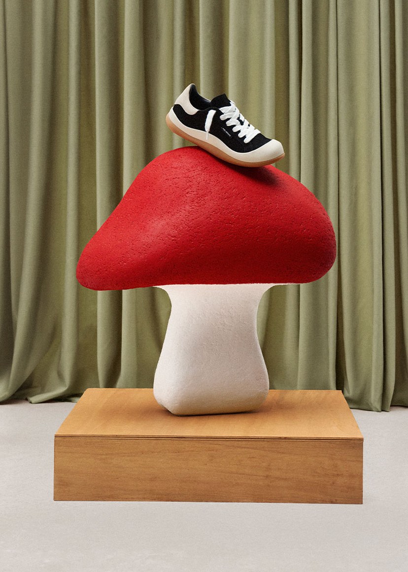 A black and white sneaker is placed on a large red and white mushroom sculpture, set on a wooden pedestal against a green curtain backdrop.