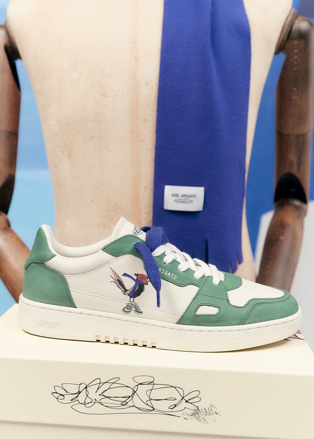 Green and white sneaker with a cartoon character, displayed on a box, in front of a mannequin draped with a blue scarf.