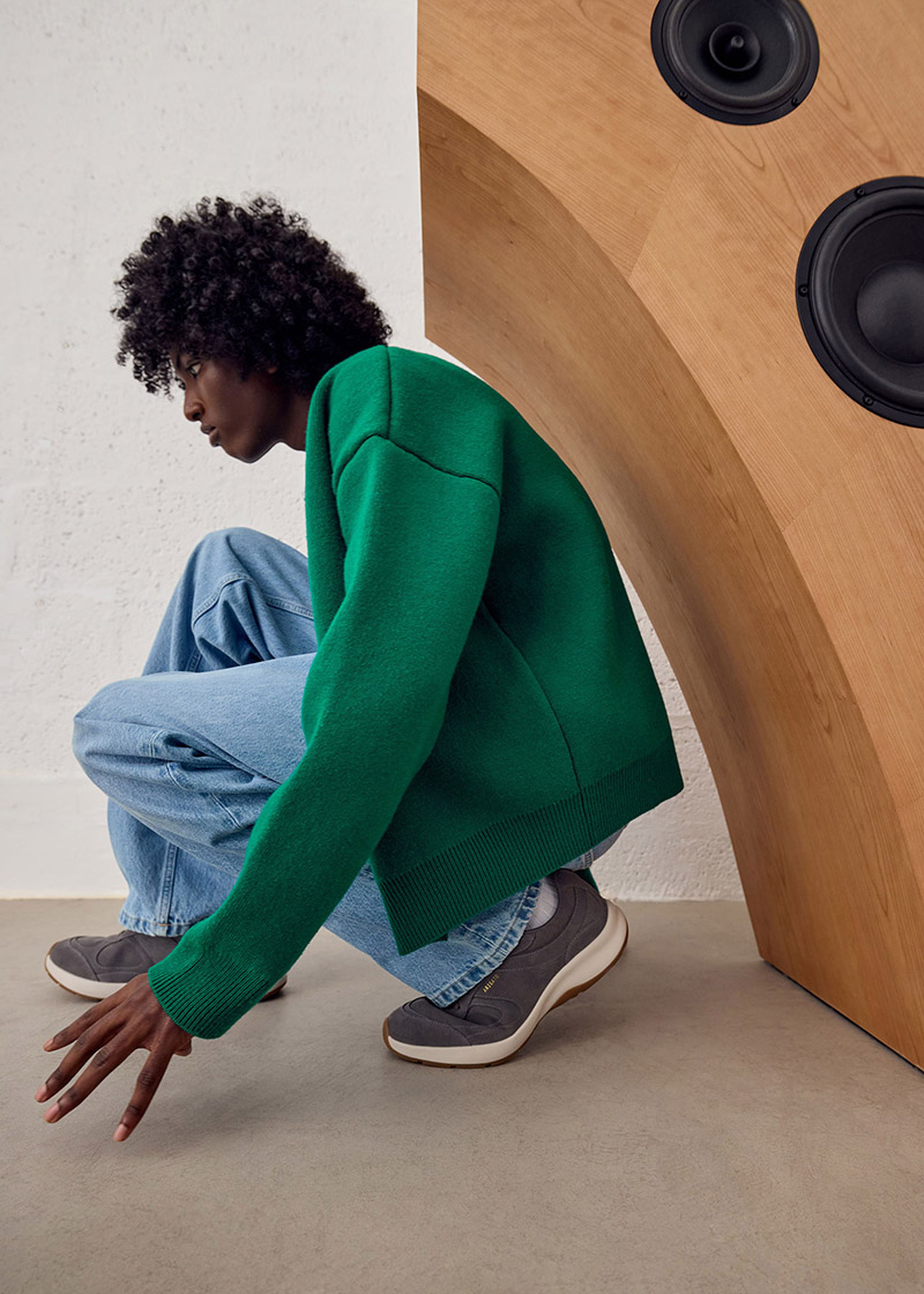 Person crouching in front of large wooden speaker, wearing green sweater, blue jeans, and gray sneakers.