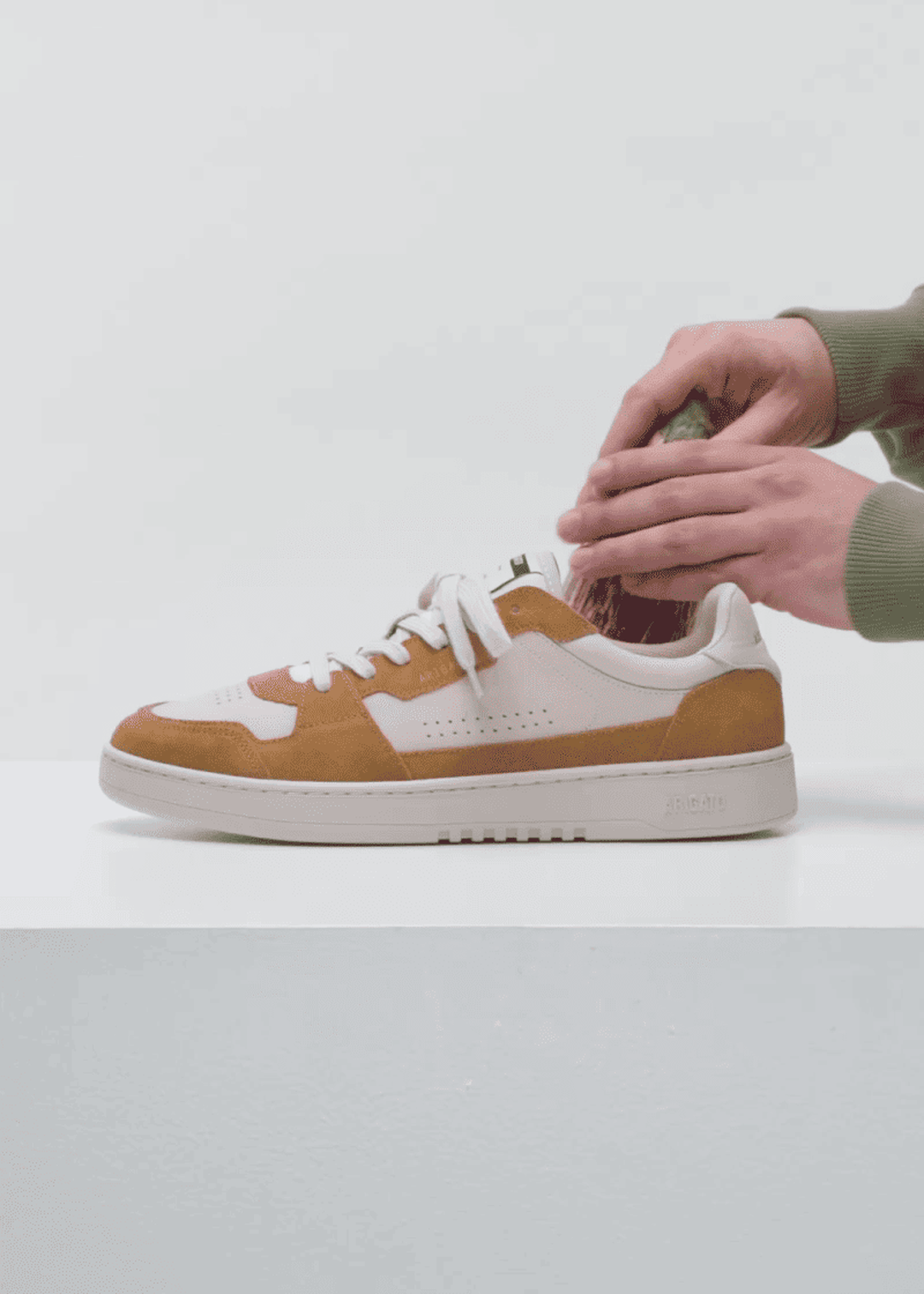 Hands brushing a white and brown sneaker with a shoe brush on a white surface, against a light gray background.