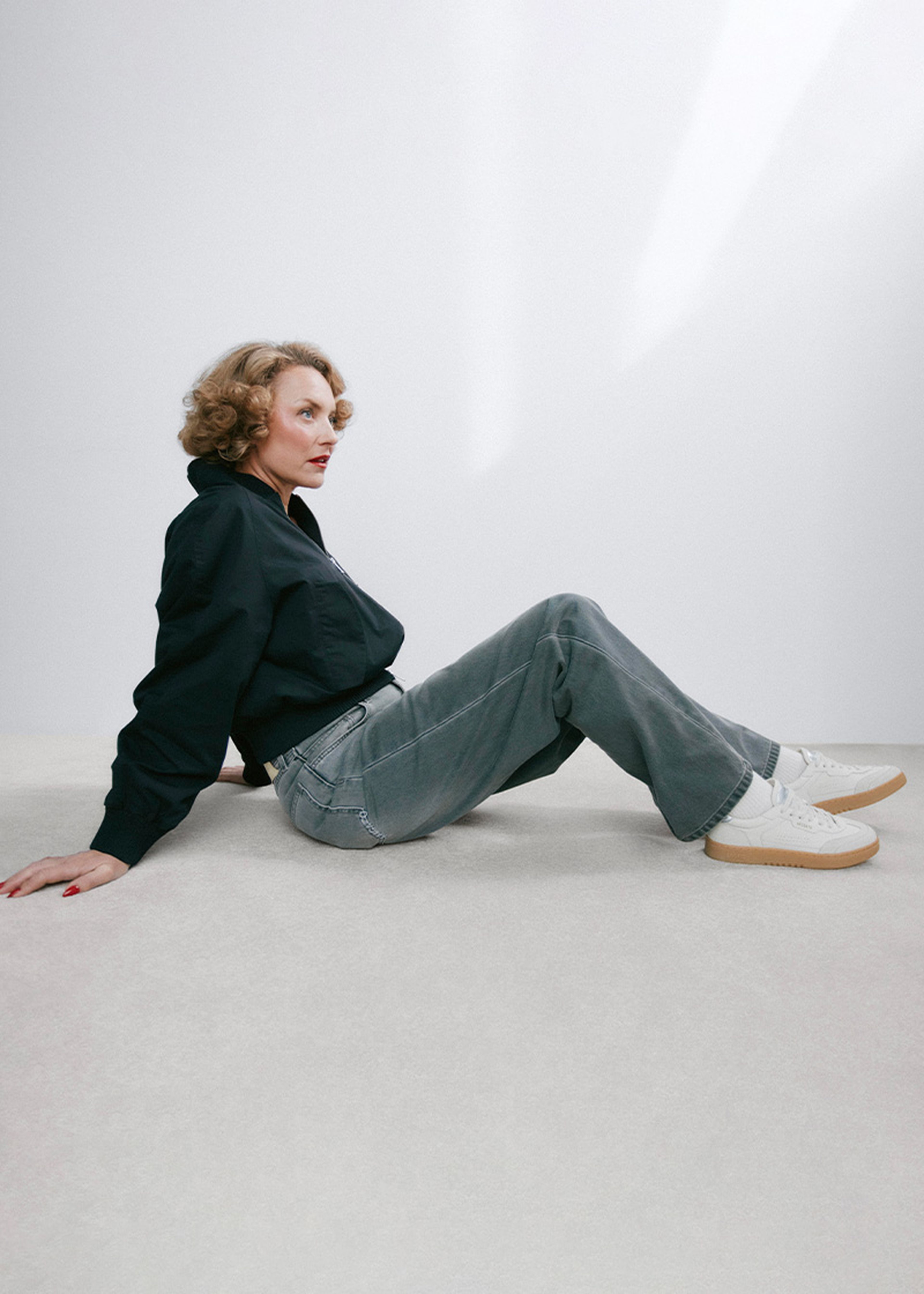 Person sitting on the floor in a black shirt and gray jeans, wearing white sneakers, against a light background.