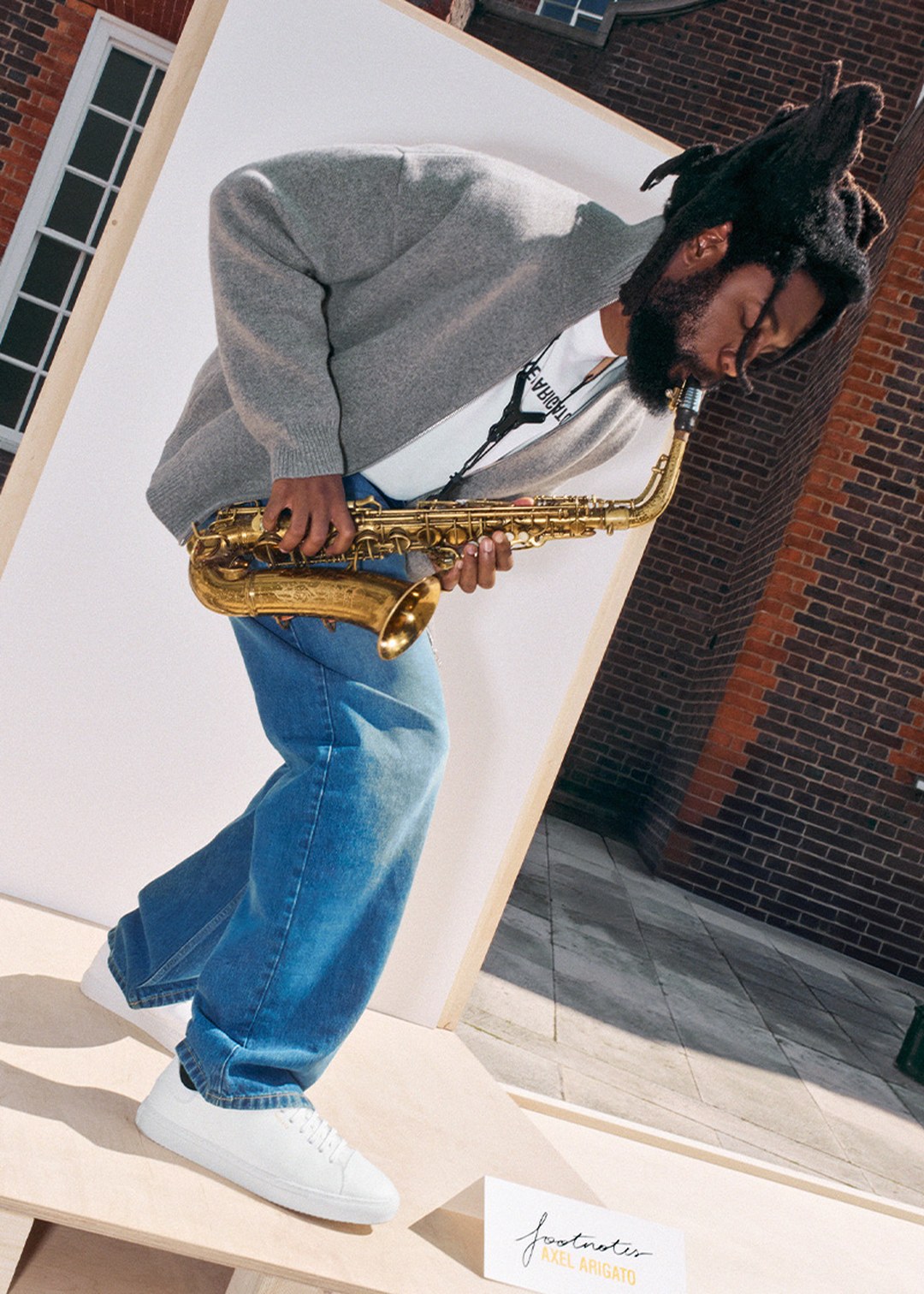 Person playing a saxophone, wearing a gray sweater, blue jeans, and white sneakers, standing on a wooden platform near a brick building.