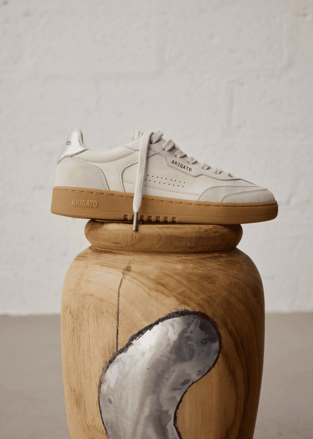 White sneaker with tan sole on carved wooden stool, textured white background.