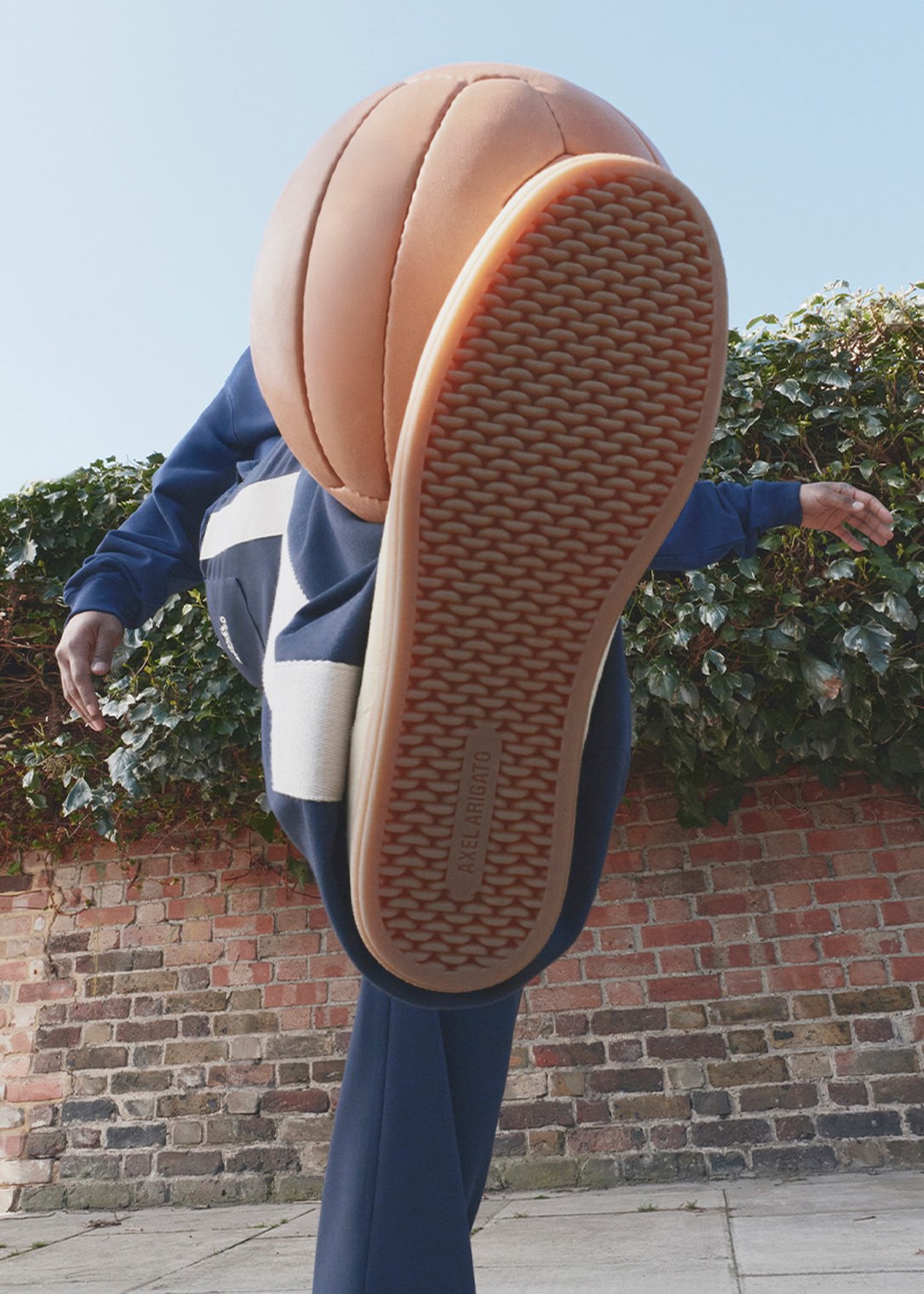 Person in a blue outfit kicking a basketball, with the shoe sole prominently visible, against a brick wall and ivy background.