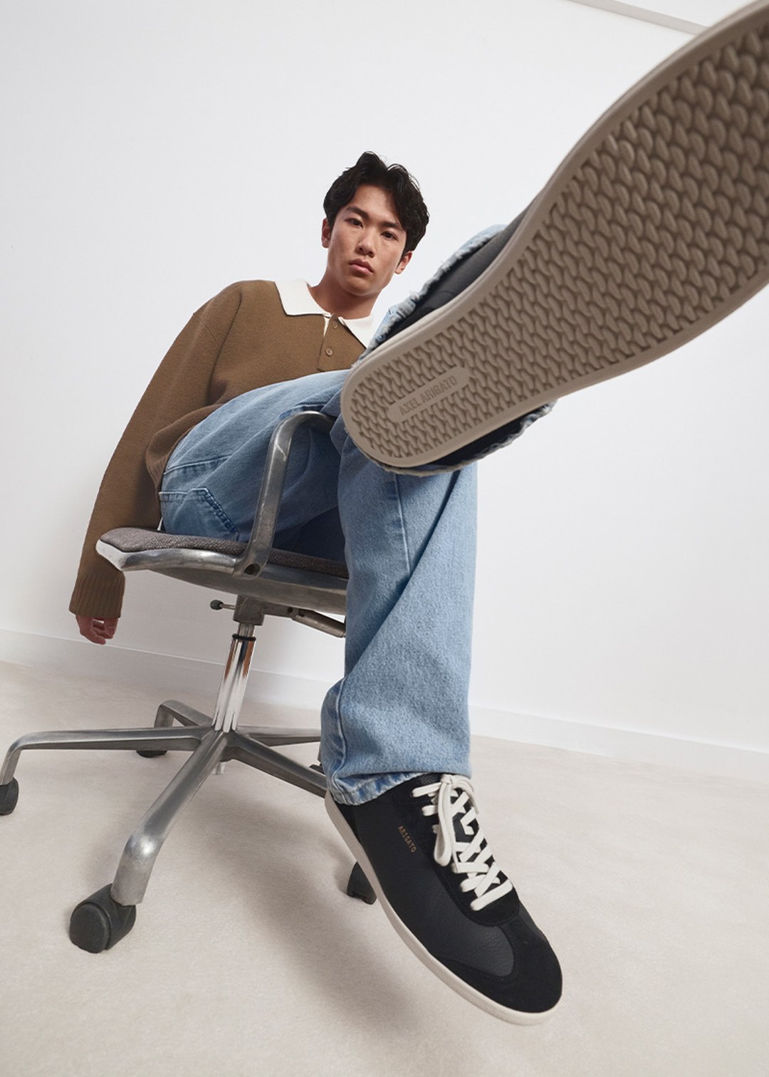 Person sitting on a wheeled chair, wearing a brown sweater, blue jeans, and black sneakers, with one foot prominently raised toward the camera.