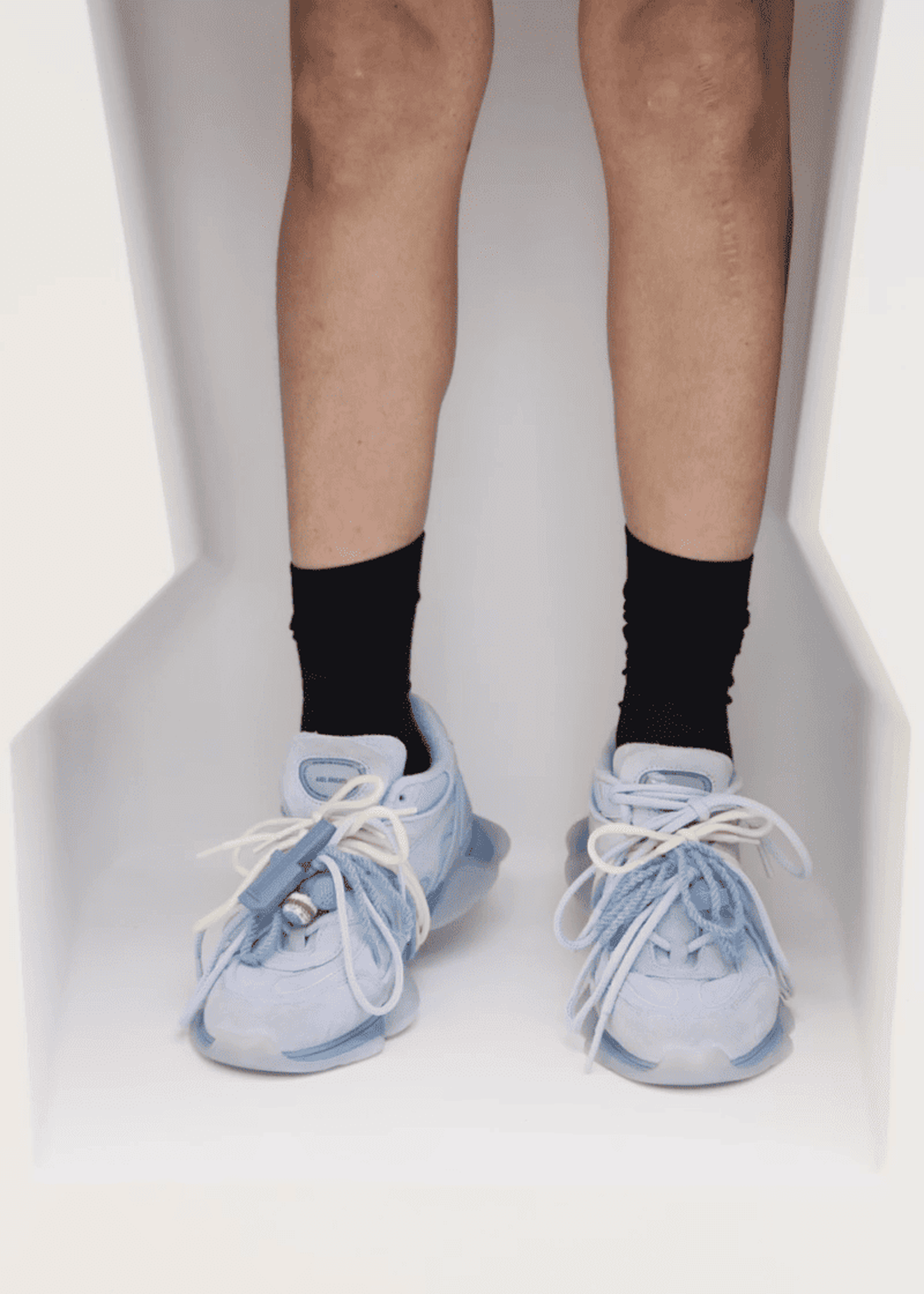 Person wearing light blue Eris sneakers with big white laces and black socks, standing in a white, angular space.