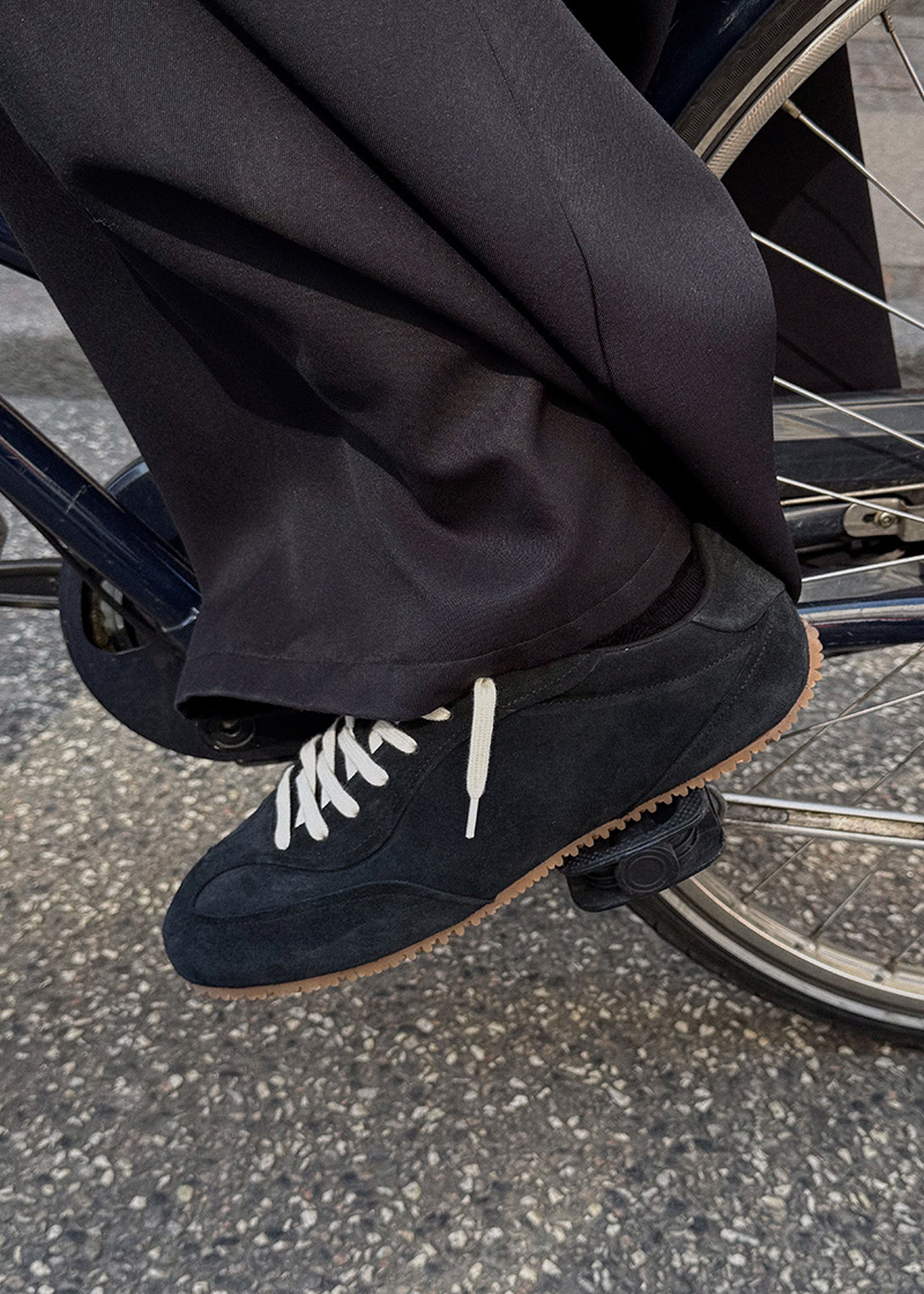 Close-up of a person wearing black pants and black sneakers with white laces, pedaling a bicycle on a textured road.