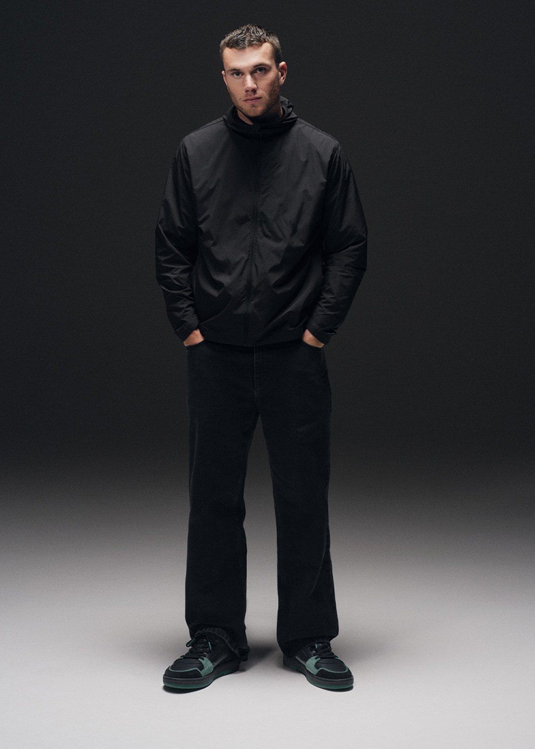 A person stands against a dark background wearing a black jacket, dark pants, and green sneakers, with hands in pockets, looking directly ahead.