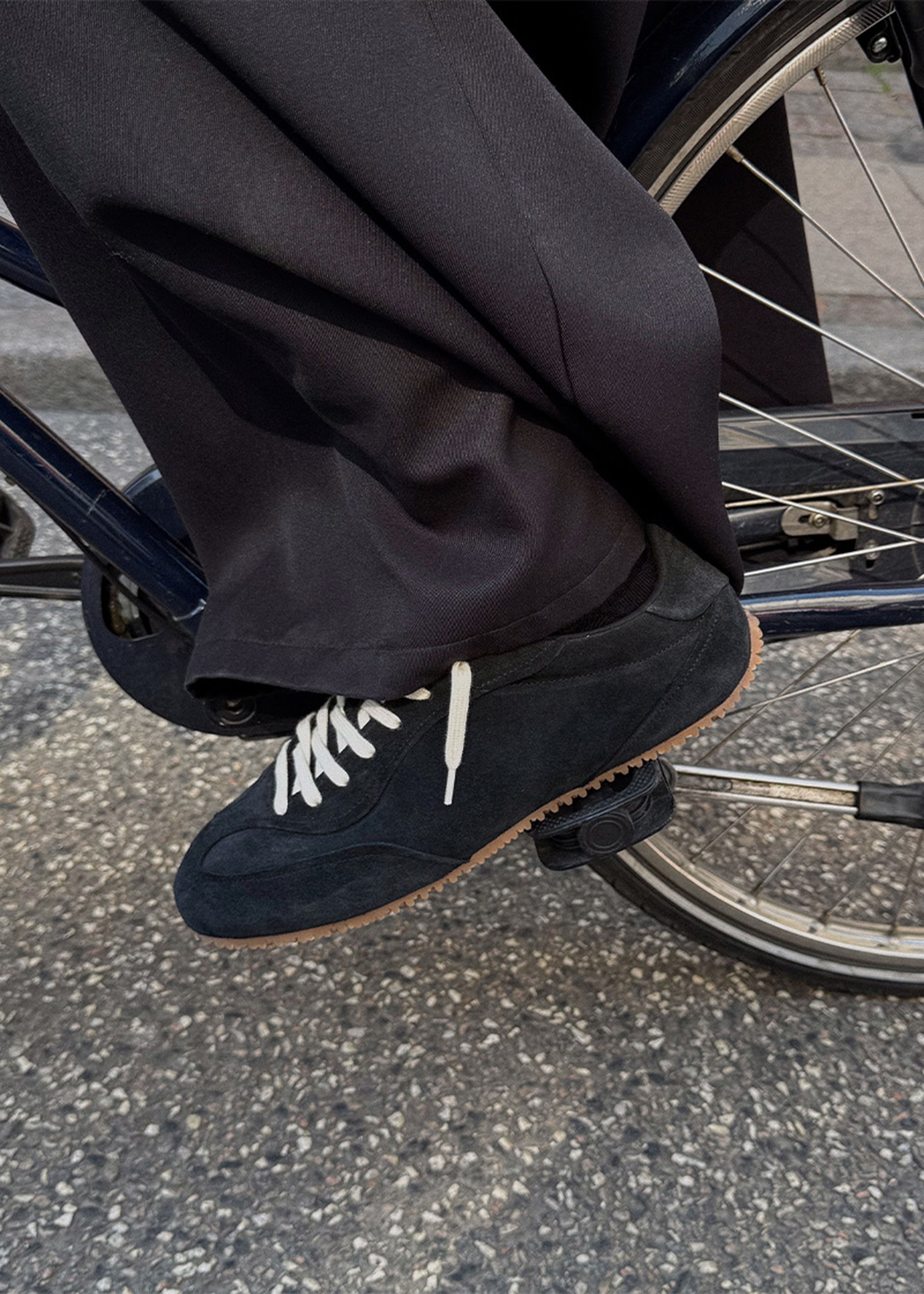 Person wearing black sneakers with white laces, resting foot on a bicycle pedal. They're dressed in dark pants.