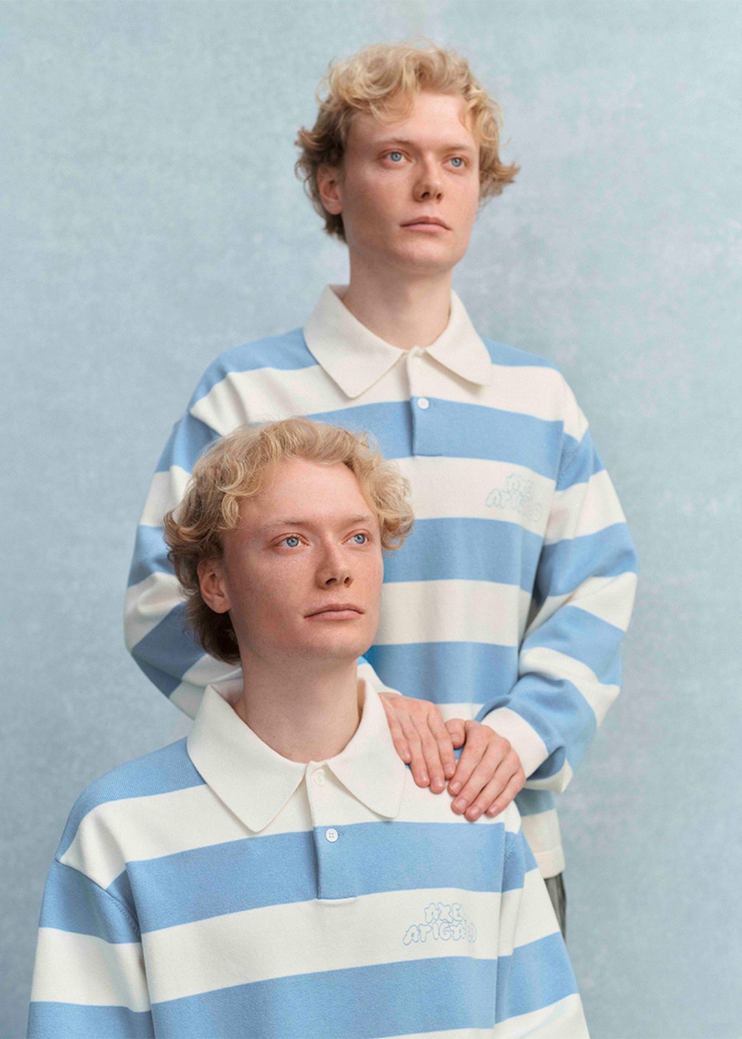 Two people with curly blond hair wear blue and white striped shirts, standing against a light blue background. One rests a hand on the other’s shoulder.