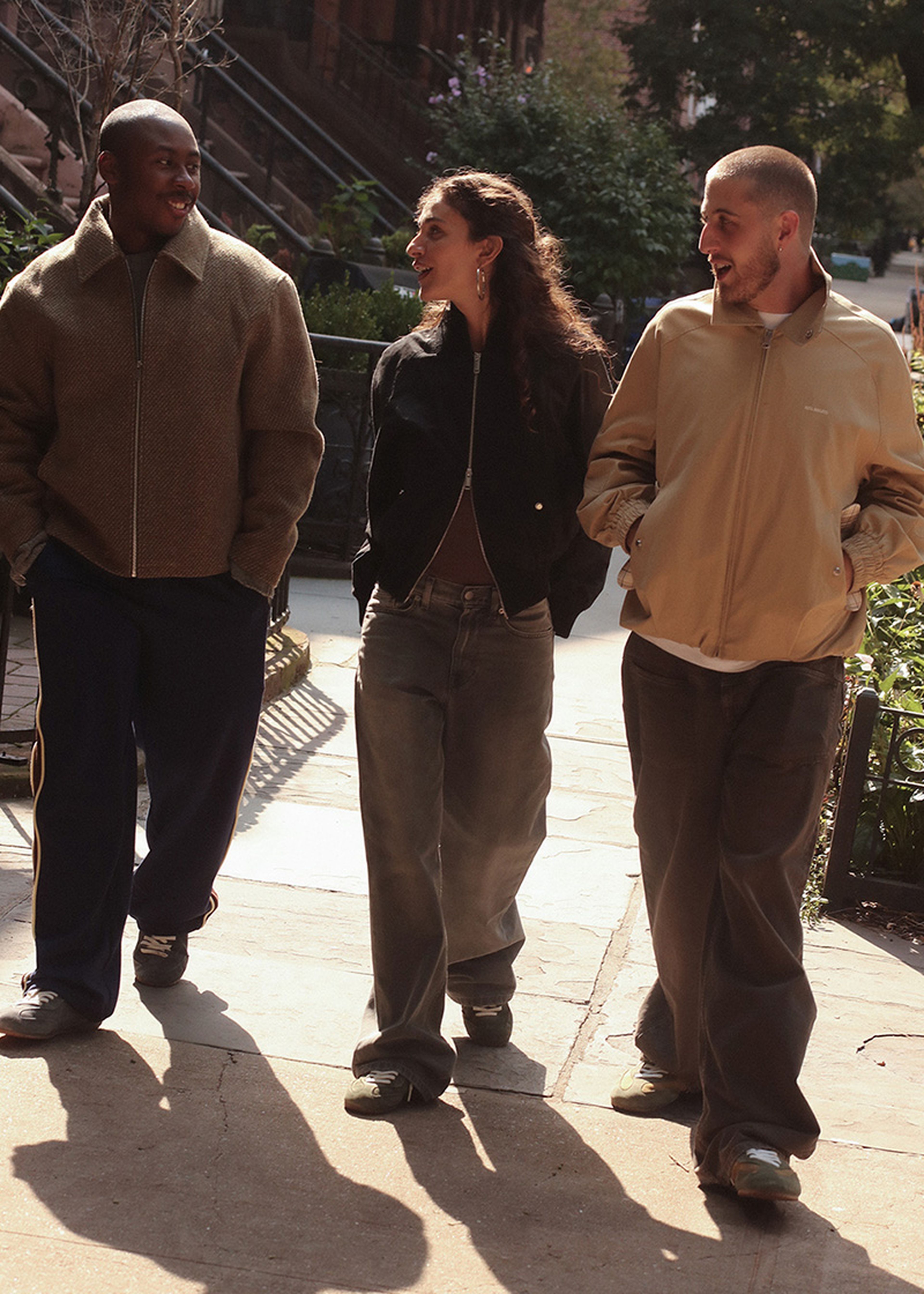 Three people walking outside on a sunny day, wearing casual jackets and jeans, smiling and engaged in conversation.