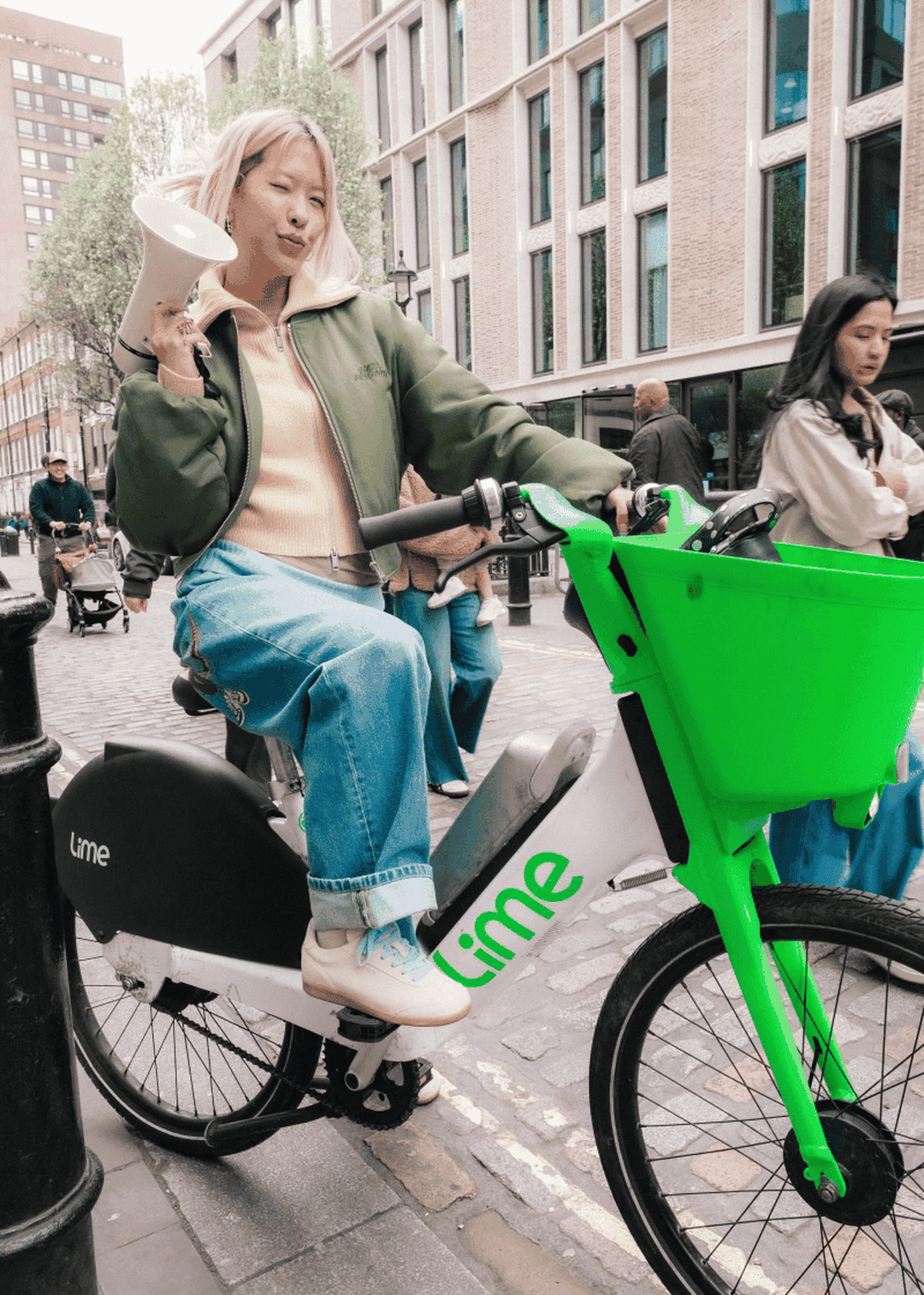 A woman poses on a Lime e-bike, holding a speakerphone, on a city street with pedestrians and buildings in the background.