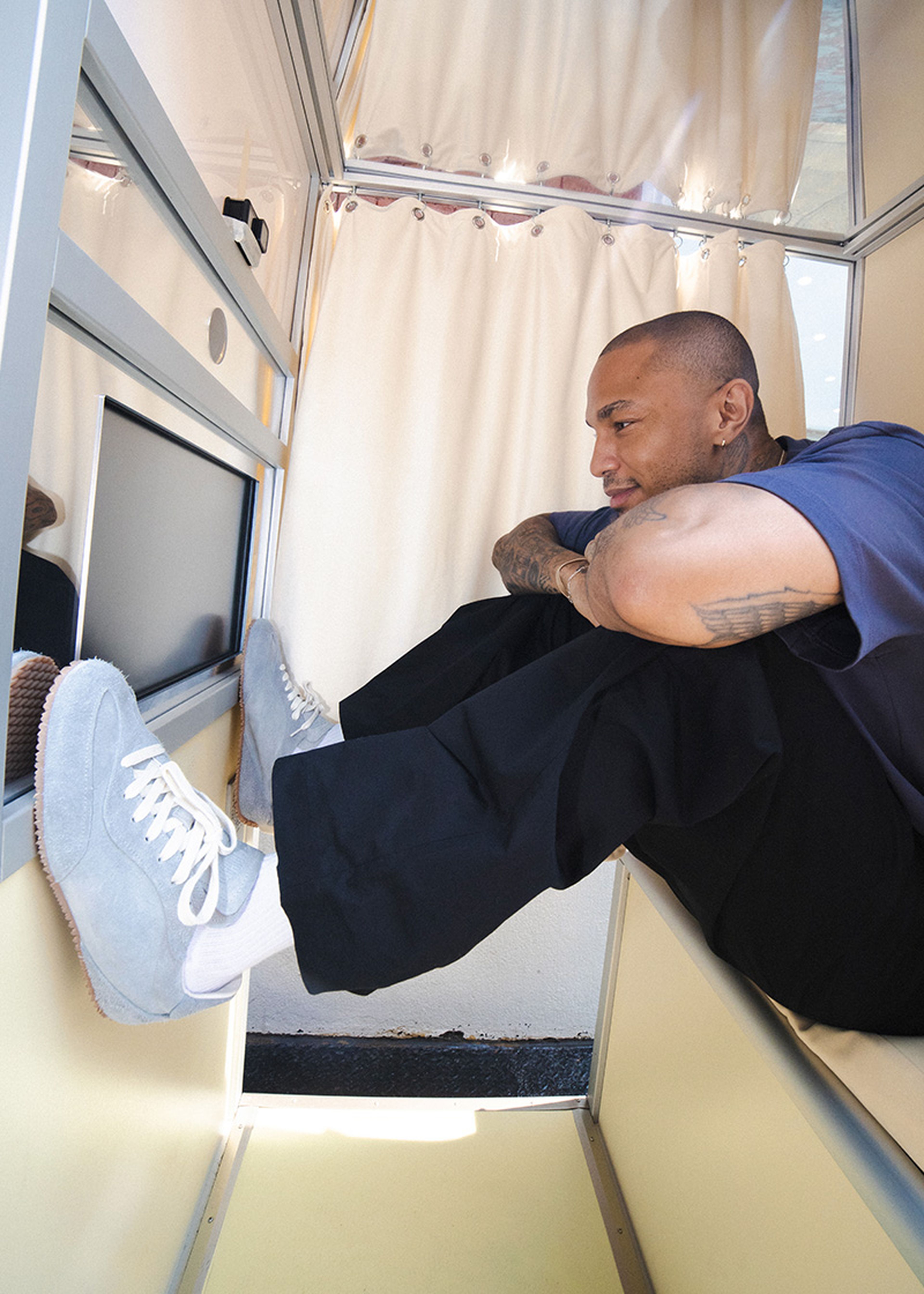 Person sitting comfortably in a small, enclosed space with light blue sneakers against a wall, resting on a cushioned bench with cream curtains.