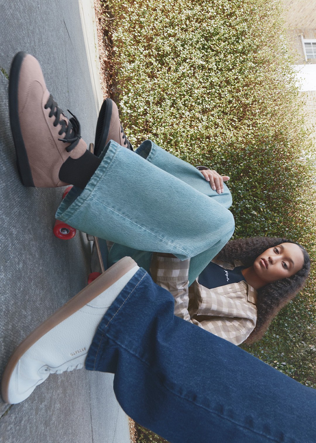 Two people on a sidewalk, one sitting on a skateboard. The person sitting wears brown shoes and jeans, while another passes by in white sneakers.