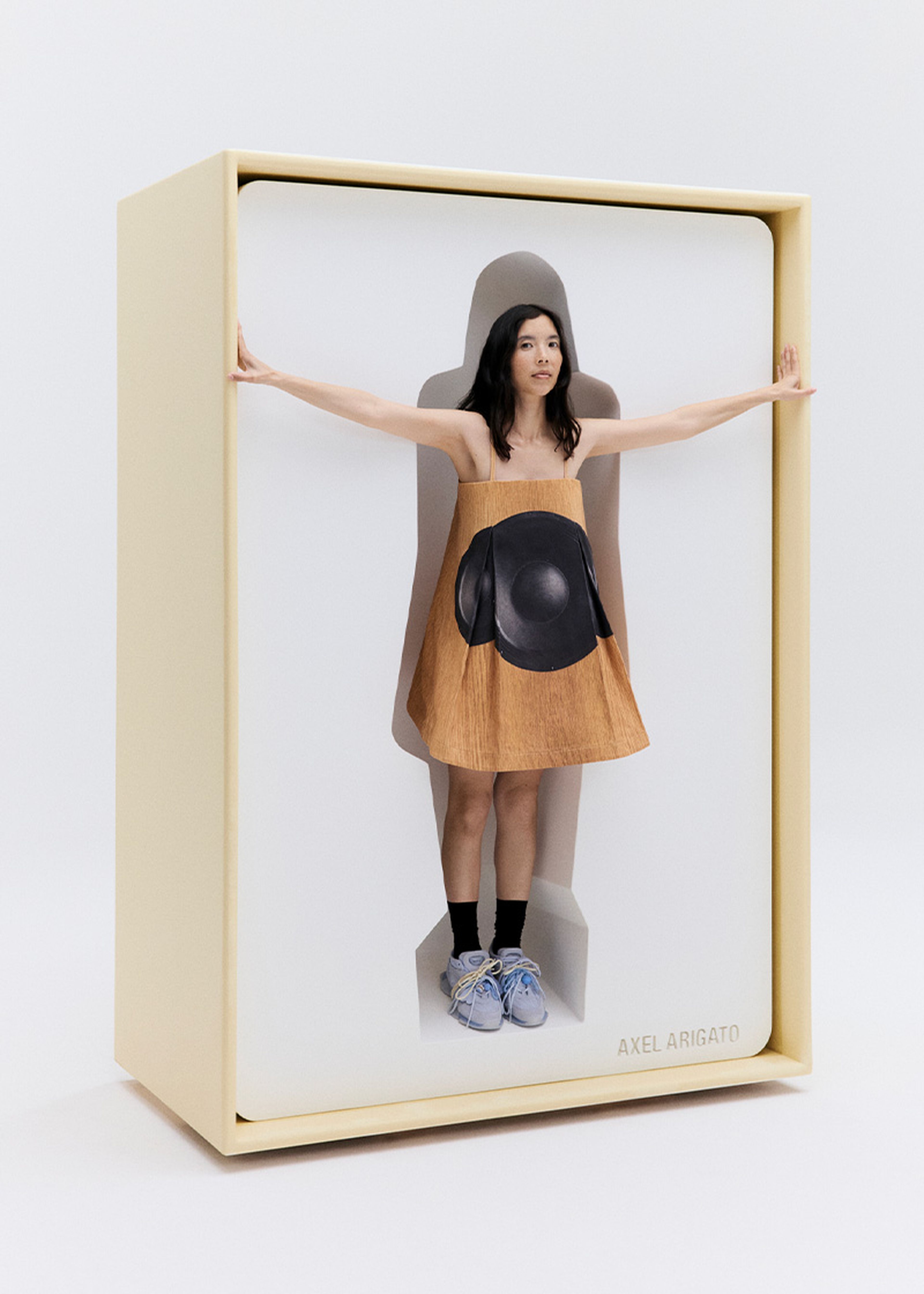 A woman stands inside a life-sized shoe box, wearing a unique dress with a black circle design, light blue Eris sneakers, and extending her arms outward.