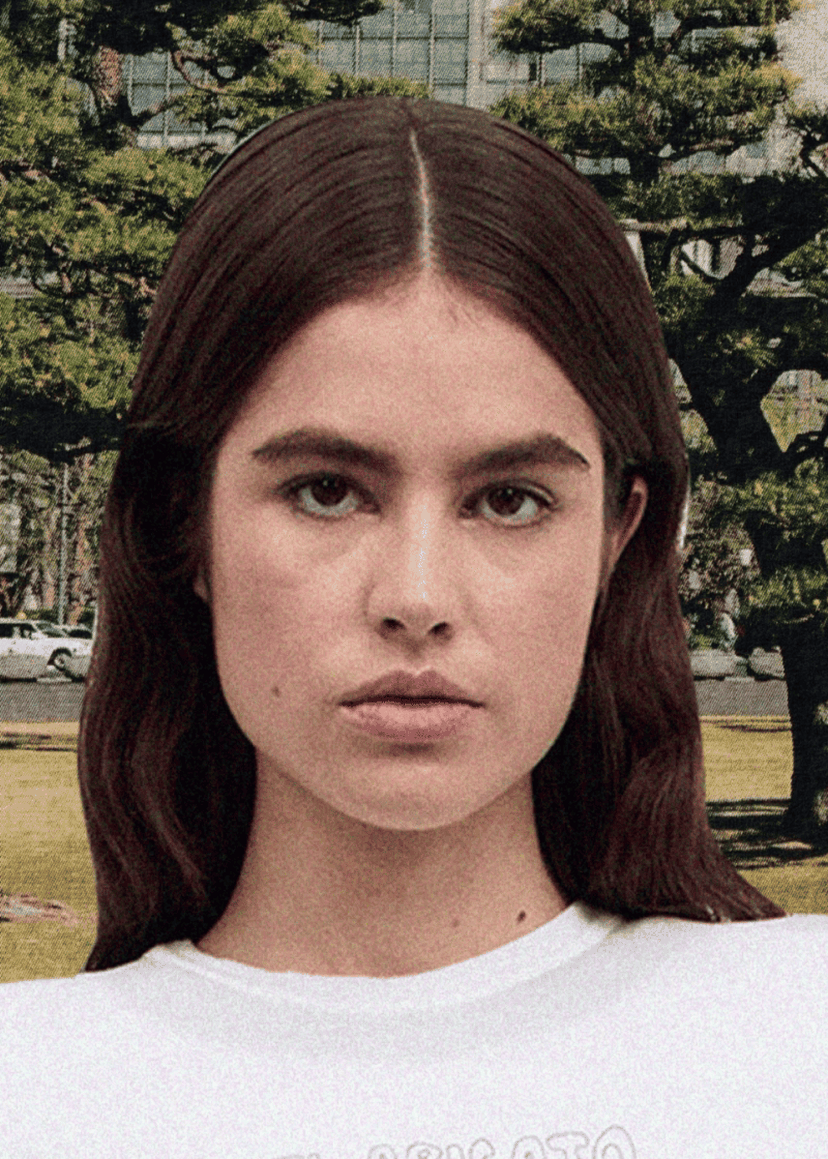 Portrait of a person with long brown hair and a white shirt standing outdoors, trees and buildings visible in the background.