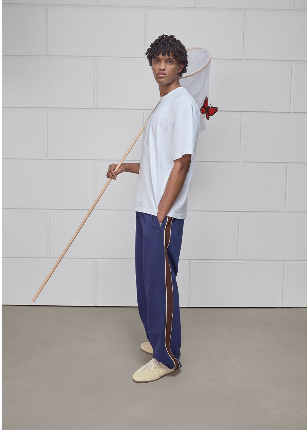 A person wearing a white shirt and blue pants holds a butterfly net with a red butterfly perched on it, standing against a tiled wall.
