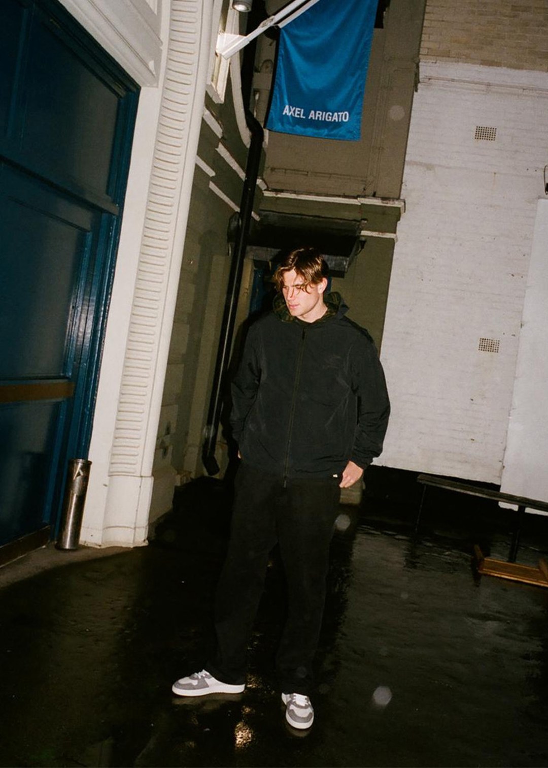 Man in black hoodie and pants stands in an alleyway at night, with a blue "Axel Arigato" flag overhead.