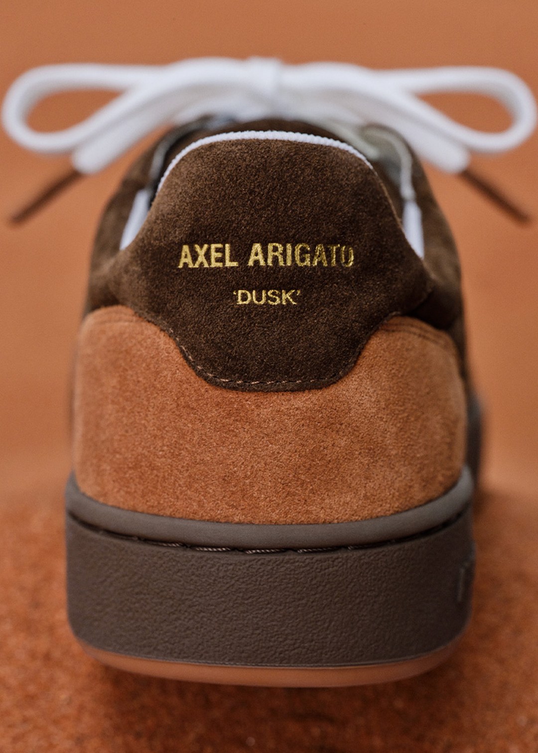Close-up of a sneaker heel with "AXEL ARIGATO" and "DUSK" in gold lettering on brown and tan suede, white laces visible in the background.
