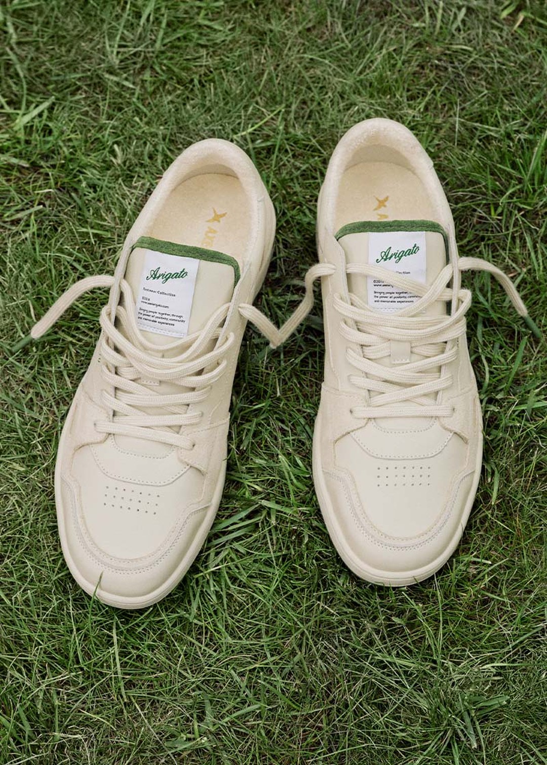 Cream-colored sneakers with green accents and logos on the tongues, laced up and placed on lush green grass.
