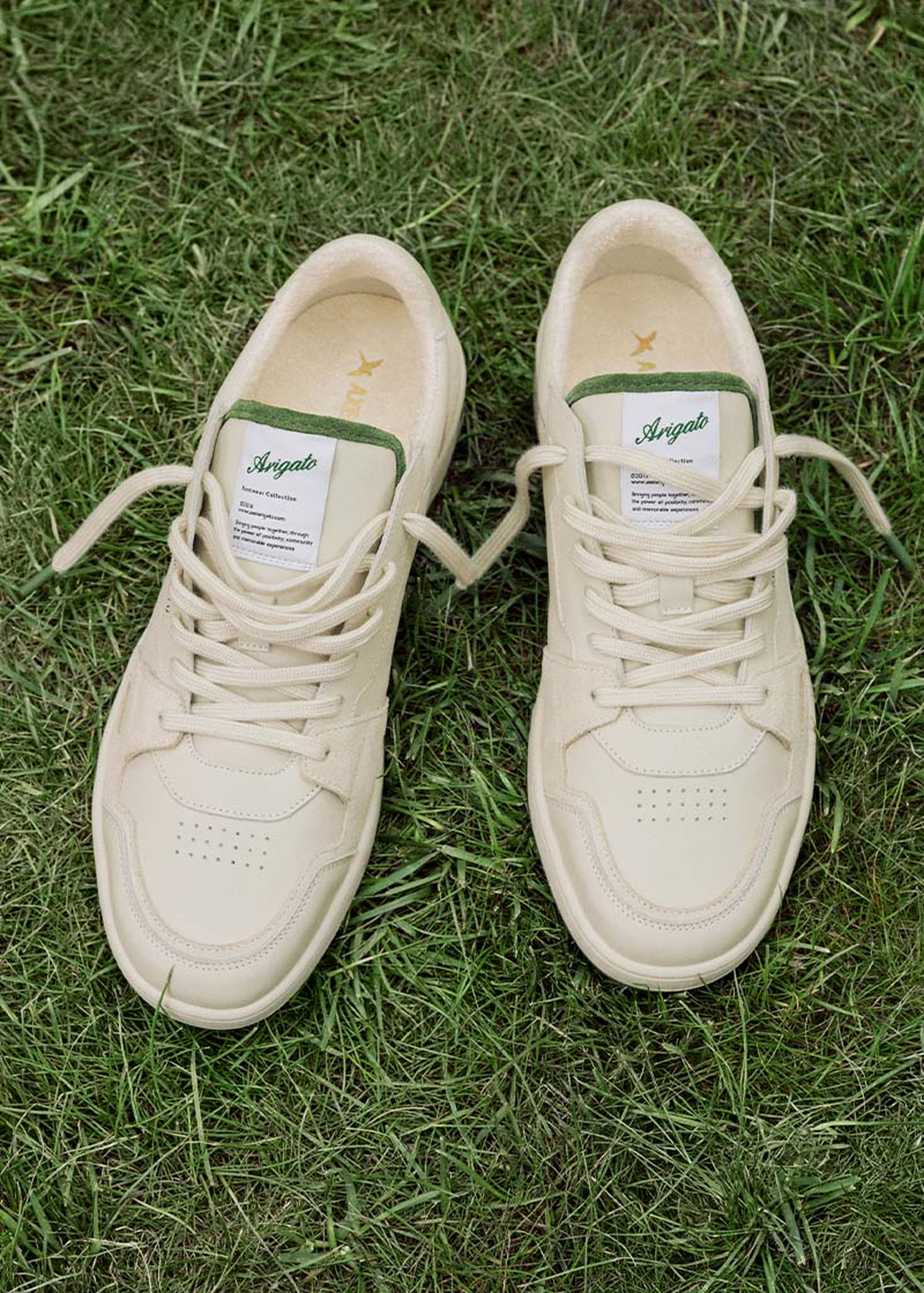 Cream-colored sneakers with green accents and logos on the tongues, laced up and placed on lush green grass.
