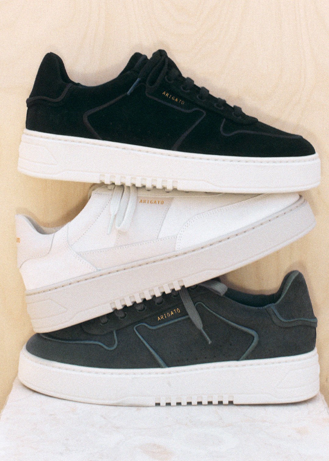 Three stacked sneakers: black, white, and gray, each with a white sole and subtle branding on a light wood background.