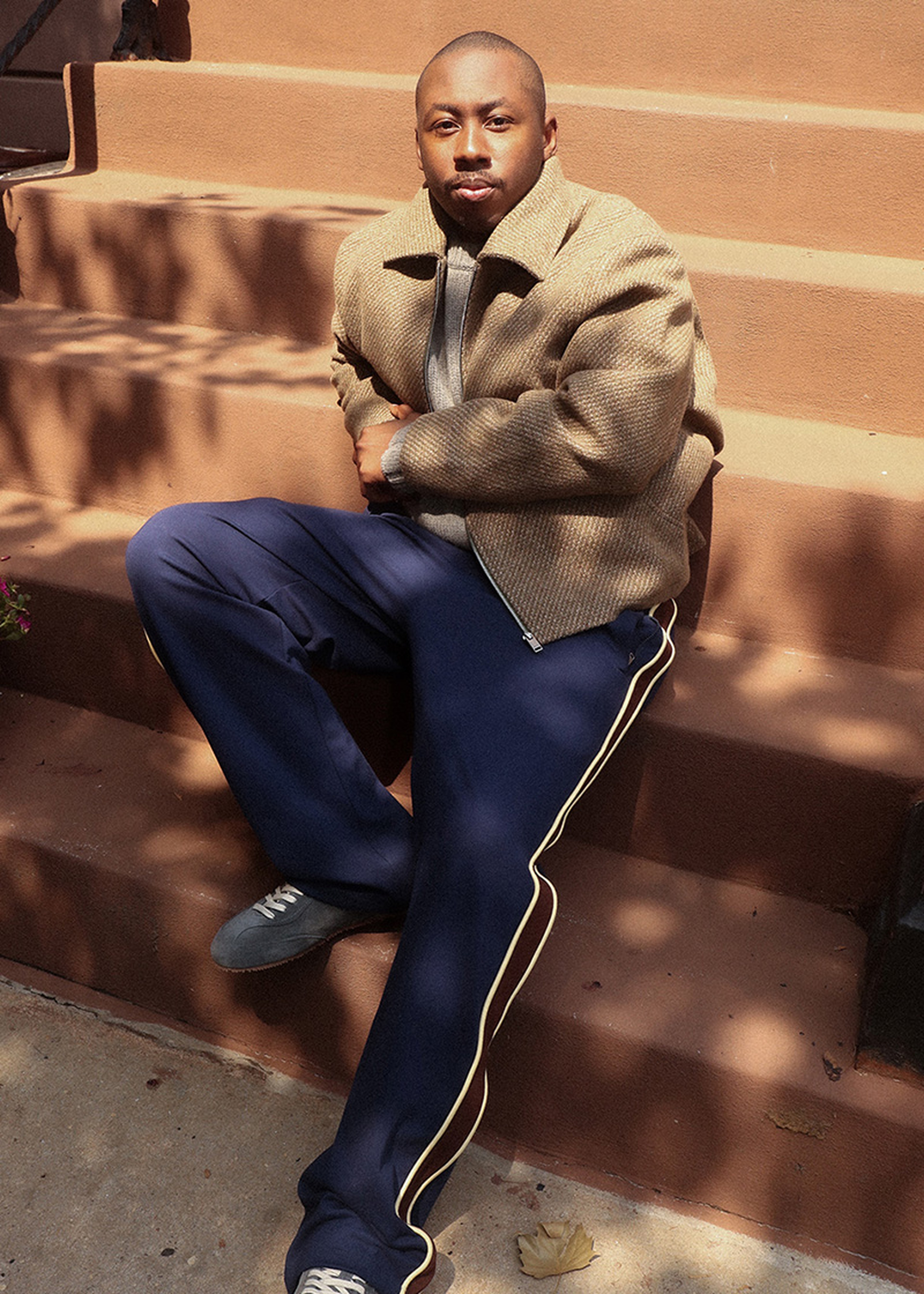Man sitting on outdoor steps, wearing a beige jacket and navy pants with stripes, looking relaxed and styled for a casual setting.
