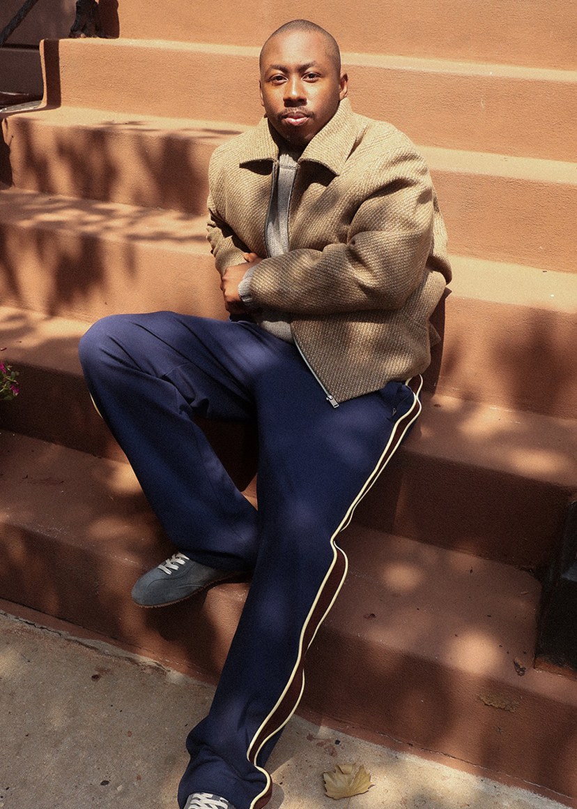 Man sitting on outdoor steps, wearing a beige jacket and navy pants with stripes, looking relaxed and styled for a casual setting.