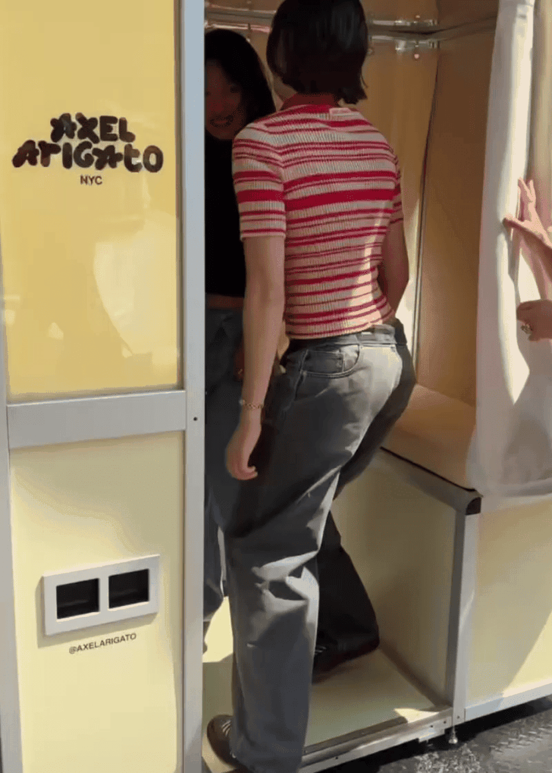 Two people in casual outfits step into a yellow photo booth. One wears a red striped shirt, and the booth displays "Axel Arigato NYC."