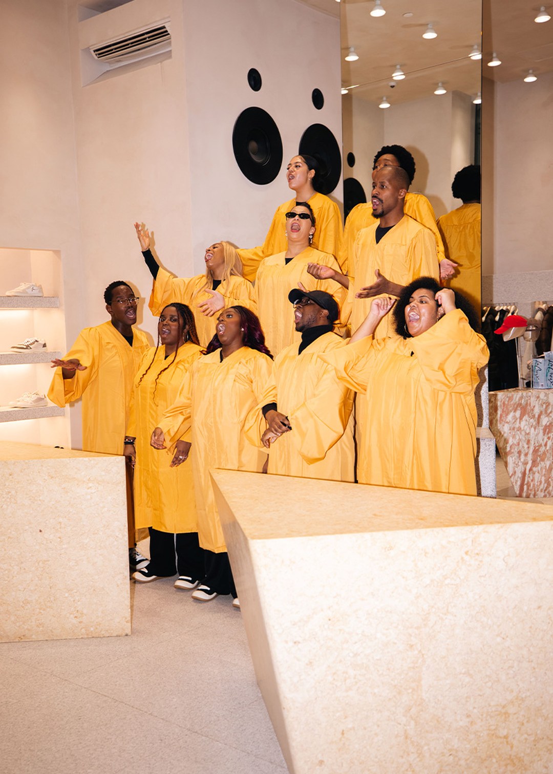 A choir in yellow robes sings energetically in a modern room with beige walls and a mirror, surrounded by shelves and speakers.