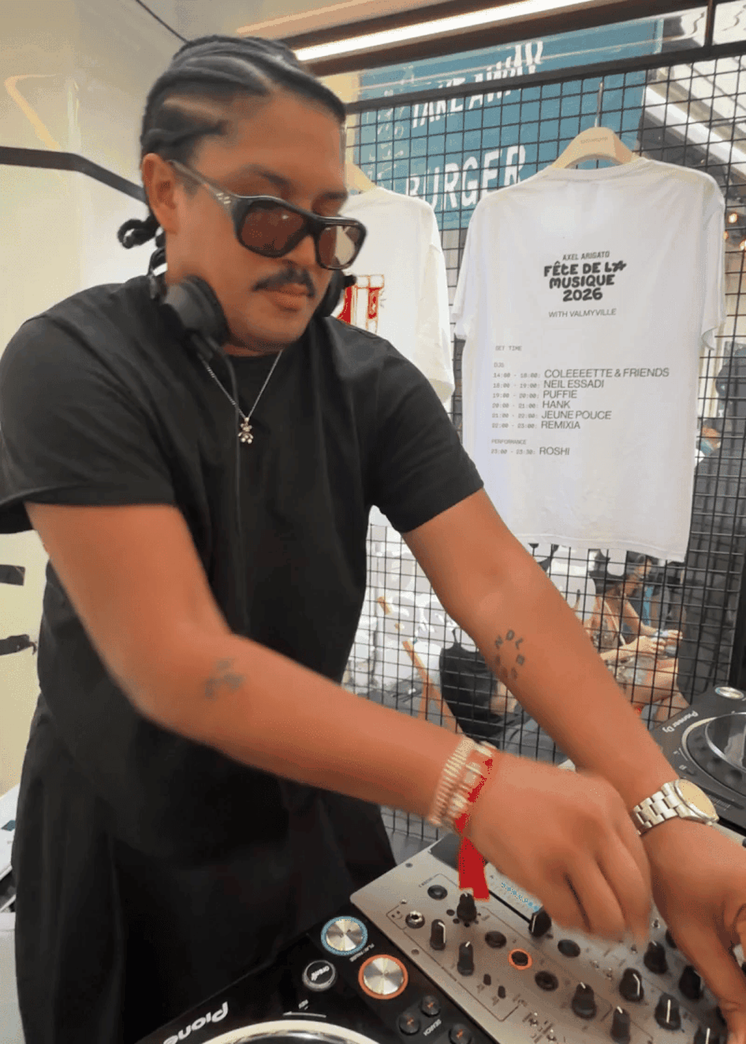 DJ wearing sunglasses and headphones, mixing music on a console. A white T-shirt with event details hangs in the background.