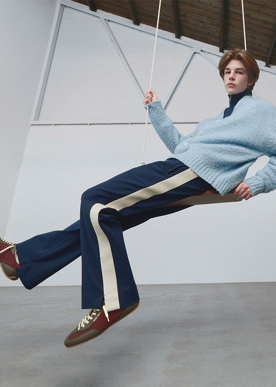 Person in a light blue sweater and navy pants with a stripe, sitting on a swing in a modern interior with high ceilings.