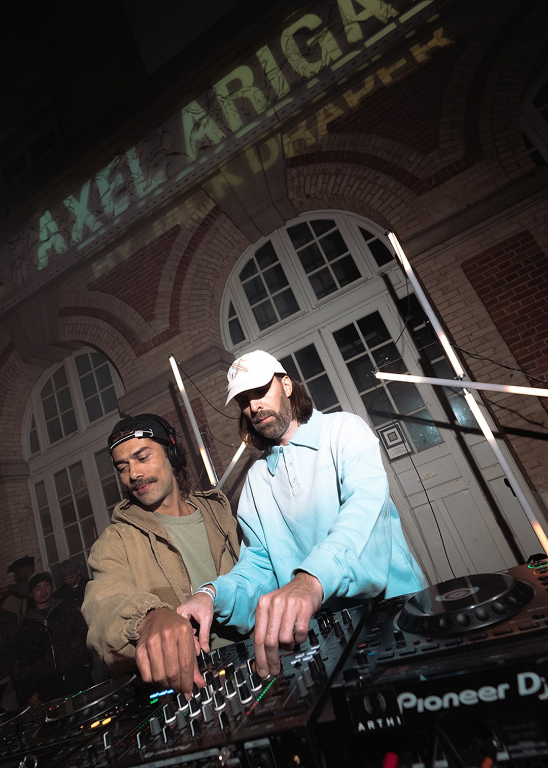 Two DJs mix tracks on a turntable at an outdoor event, with "Axel Arigato" projected on a building behind them.