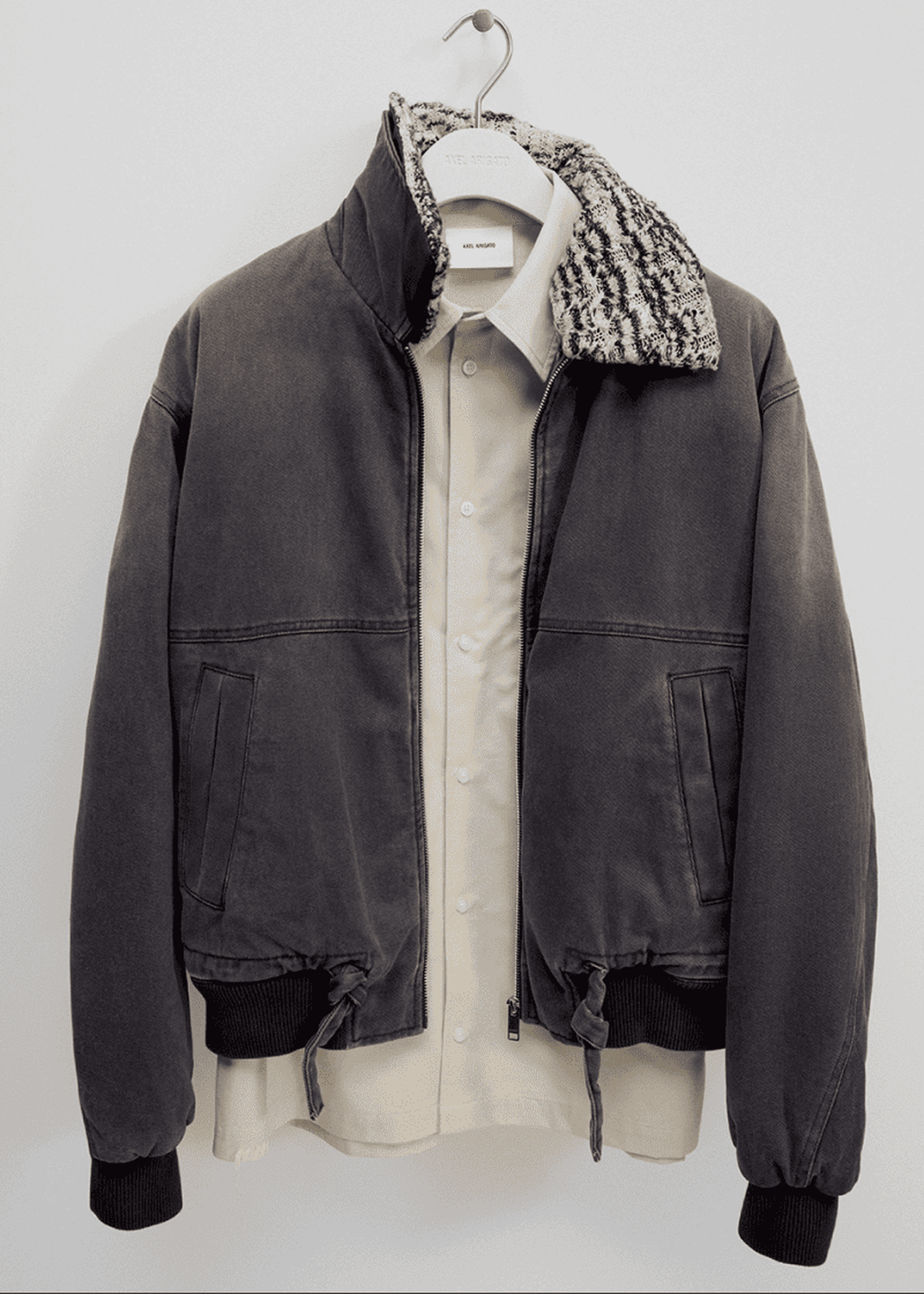 Dark bomber jacket with a textured collar, layered over a beige button-up shirt on a hanger against a white wall.