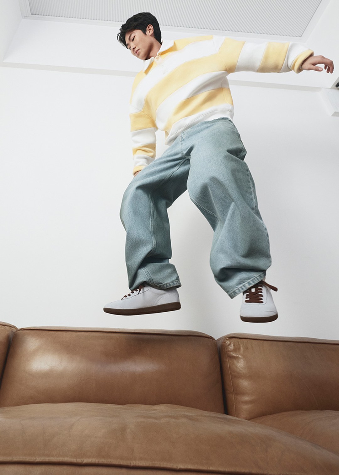 Man in a yellow and white striped sweater and jeans jumping on a brown leather couch.