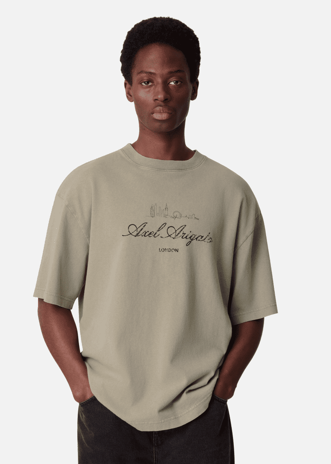 A person wearing a beige T-shirt with "Axel Arigato" and a city skyline design. Hands in pockets, looking forward against a white background.