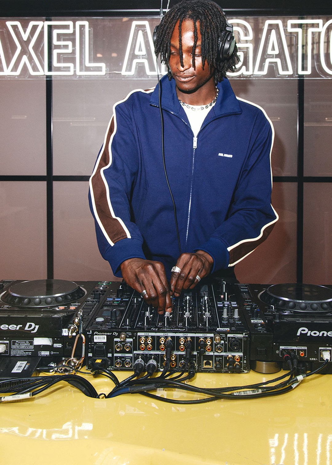 DJ in a blue track jacket mixing music on a Pioneer DJ controller with headphones, standing in front of a sign with "AXEL ARIGATO".