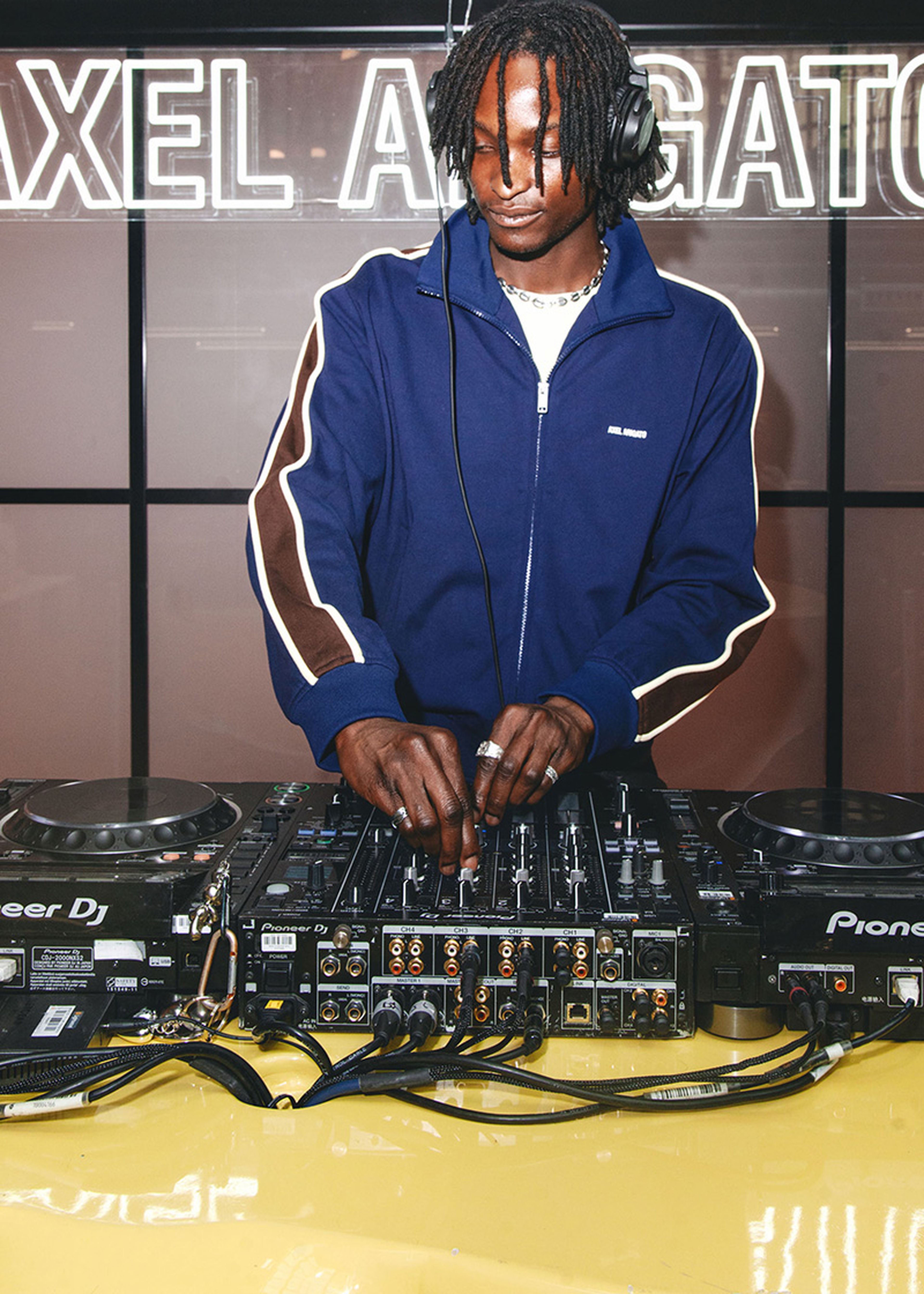 DJ in a blue track jacket mixing music on a Pioneer DJ controller with headphones, standing in front of a sign with "AXEL ARIGATO".