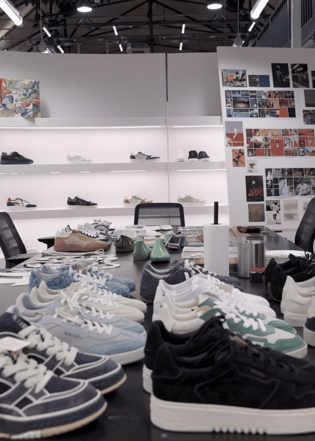 Sneaker design studio with various shoe prototypes on a table, displayed shelves in the background, and a mood board on the right wall.